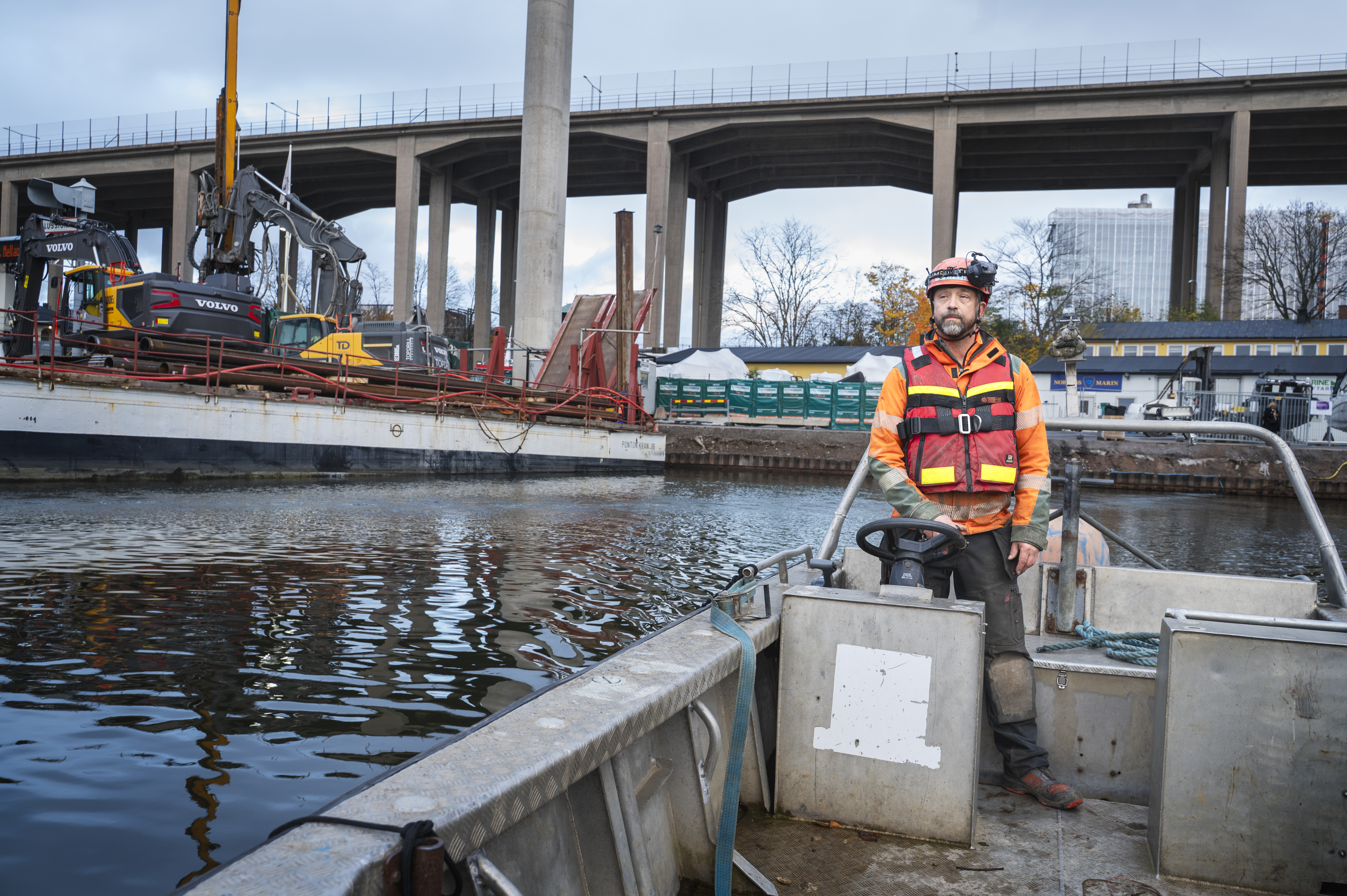 En byggarbetare med skyddsutrustning står på en liten båt i en flod nära en byggarbetsplats med maskiner och en viadukt.