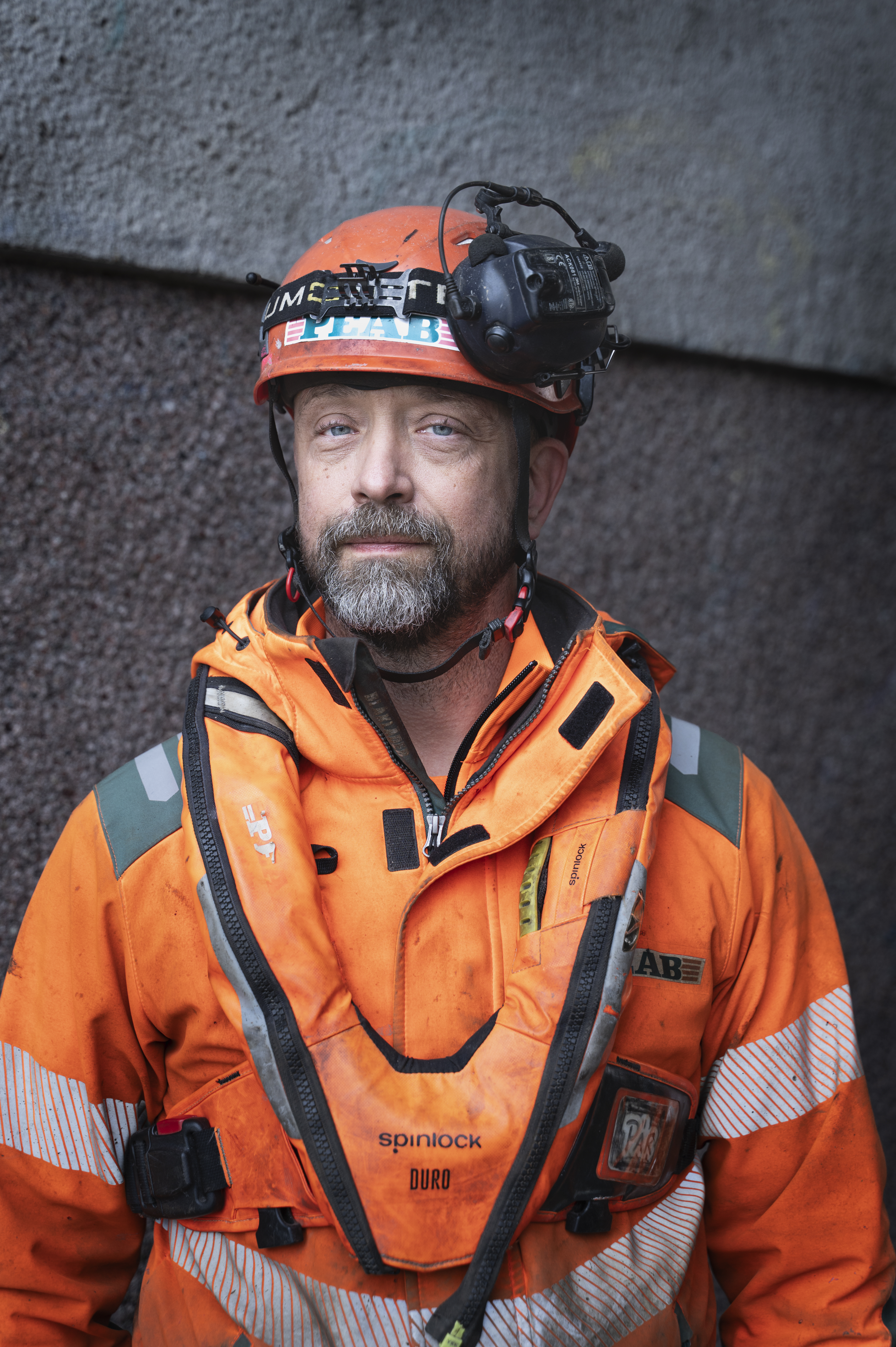 En man med orange säkerhetsjacka, hjälm med pannlampa och sele står framför en vägg och tittar mot kameran.