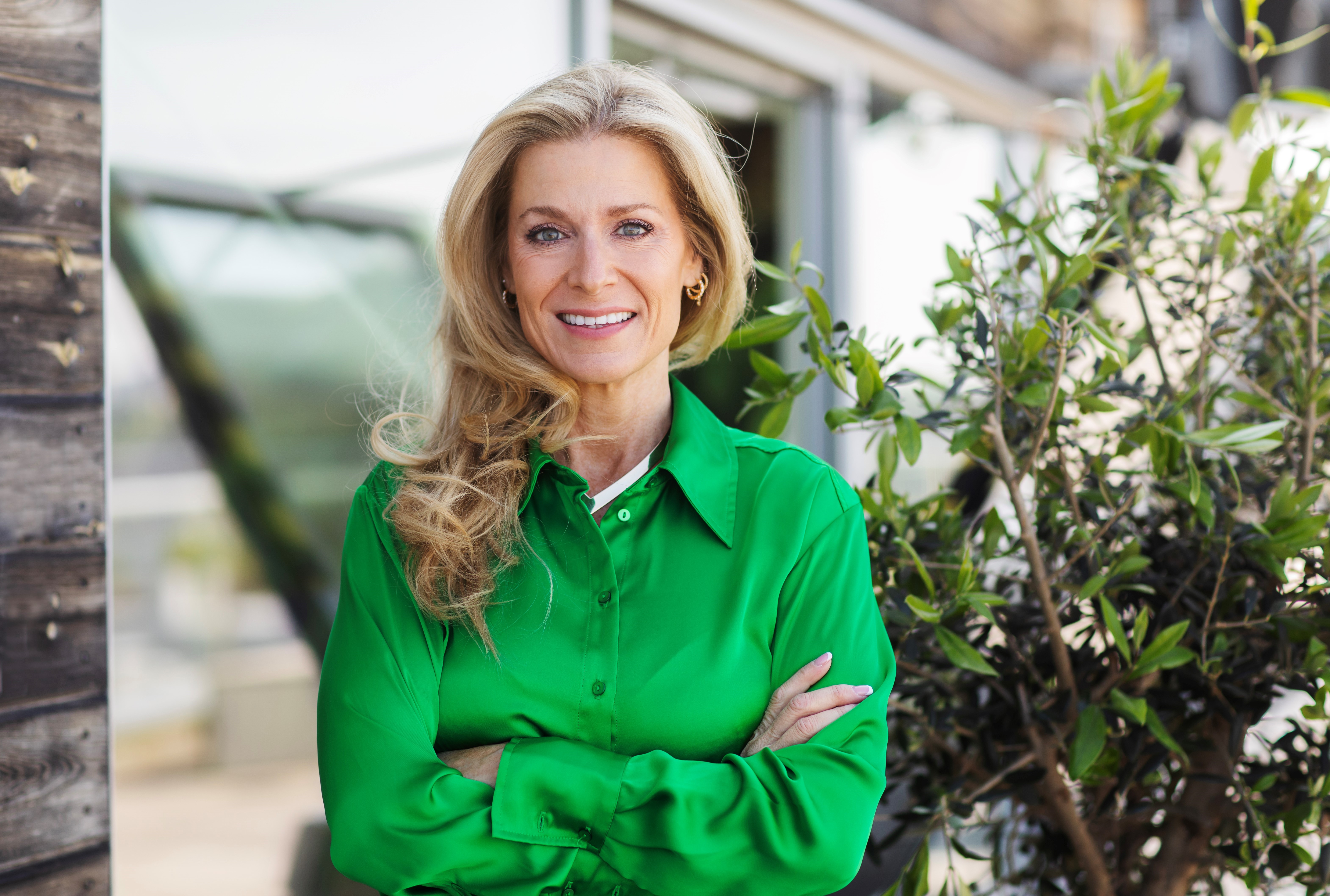 En kvinna med långt blont hår och grön blus står utomhus med armarna i kors och ler framför växter och en byggnad.