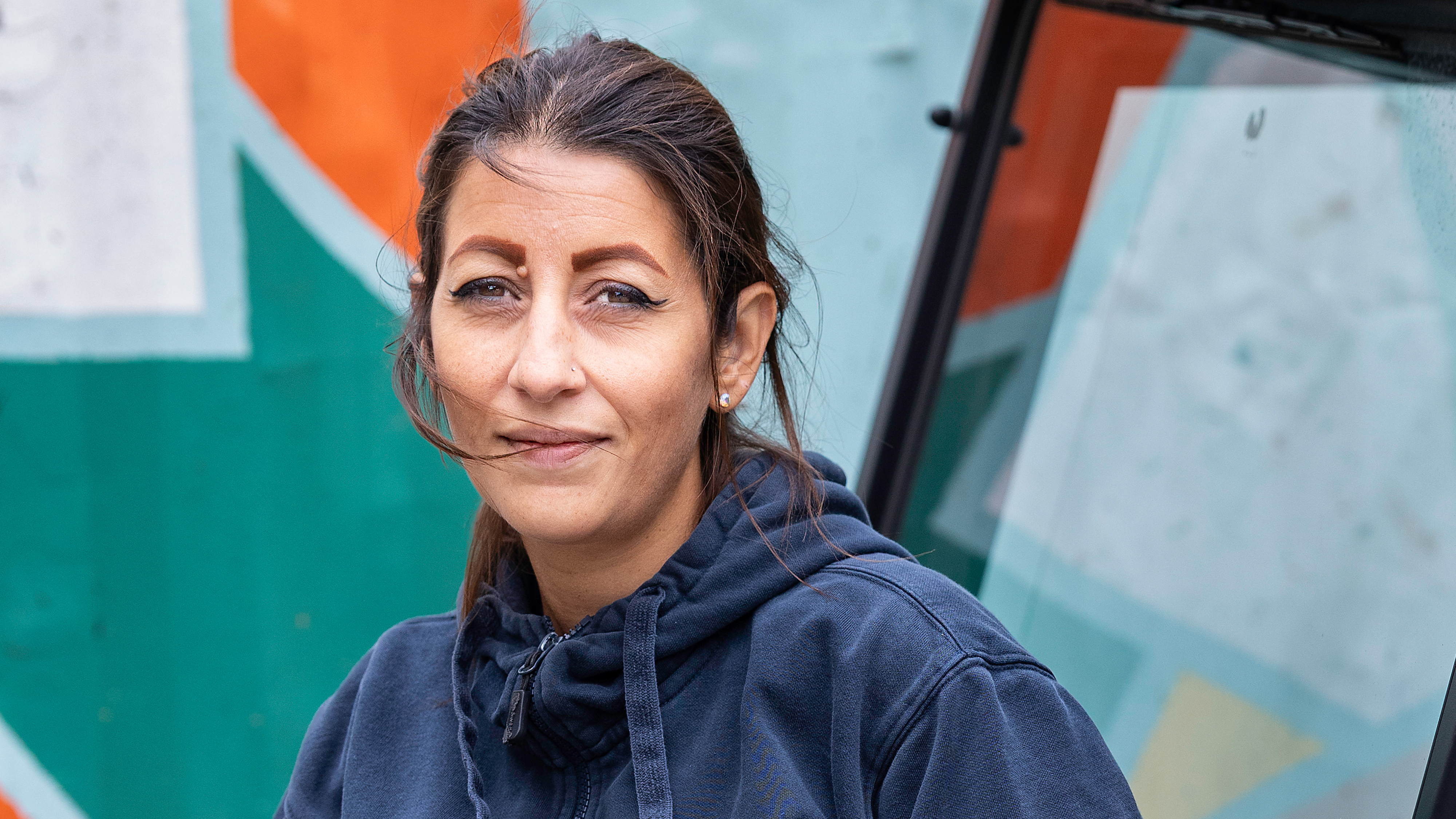 En kvinna med brunt hår i hästsvans och blå huvtröja står utomhus framför en muralmålning.