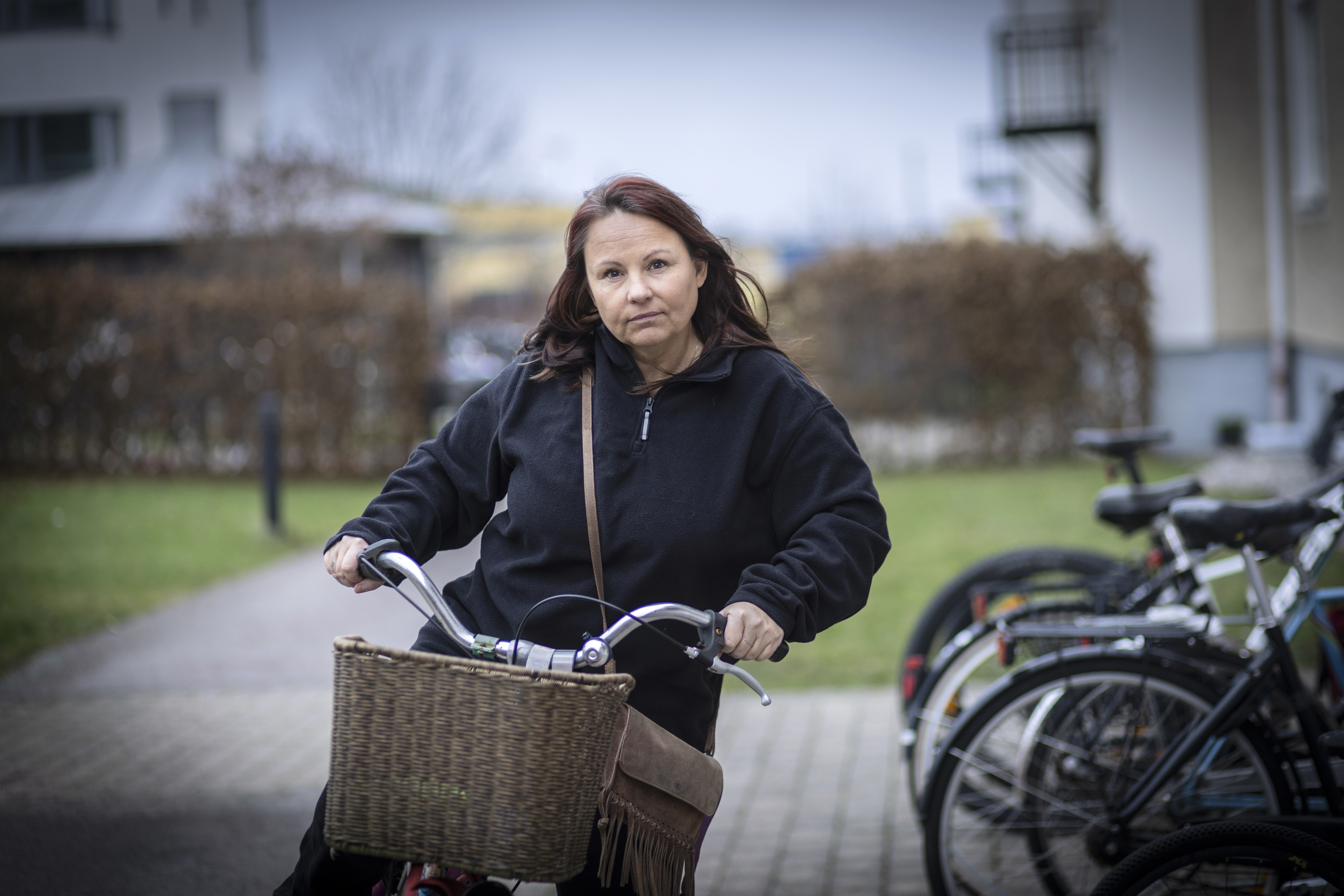 Jessica Axelsson med svart jacka cyklar på en asfalterad väg med en cykelkorg, nära parkerade cyklar och byggnader.