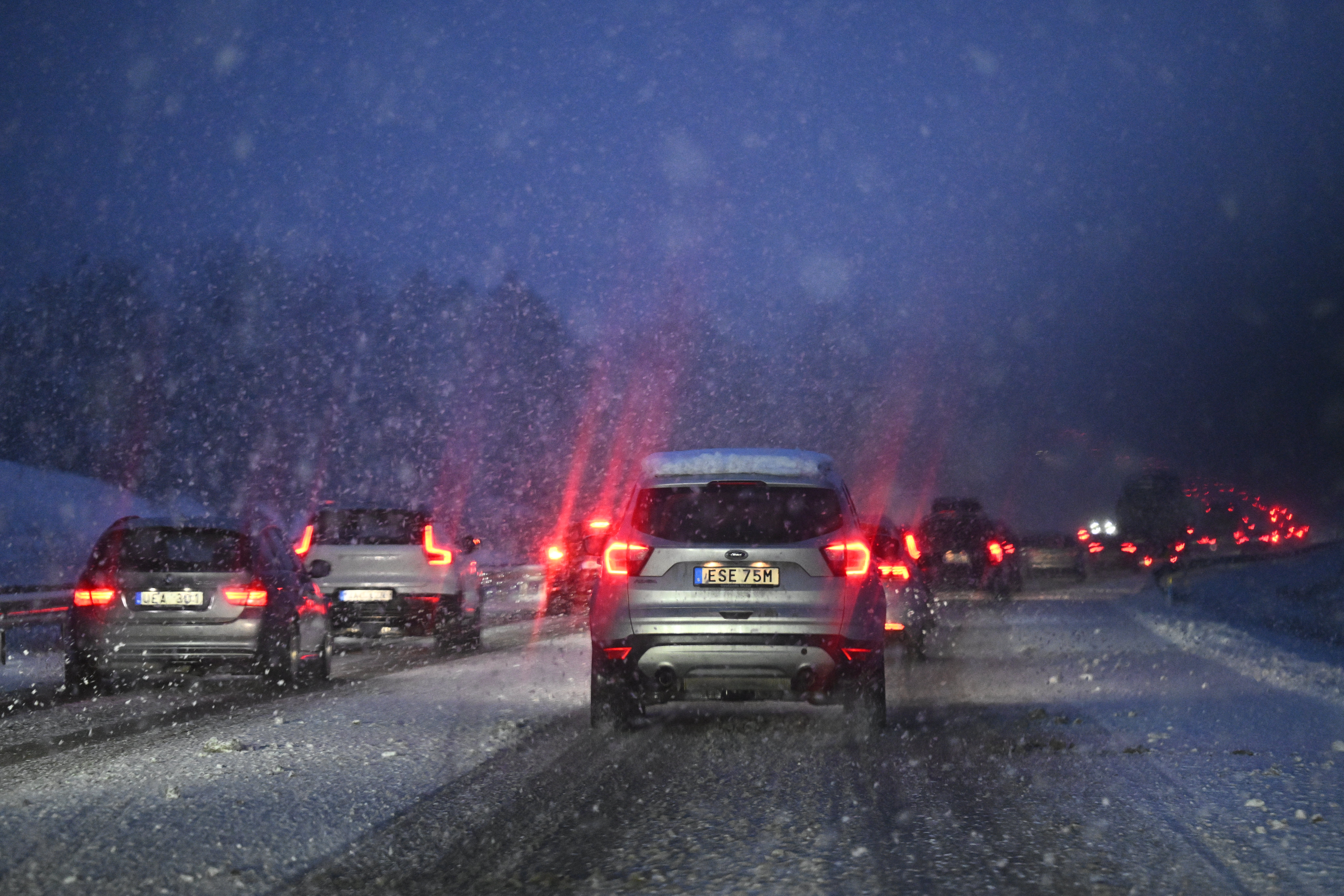 Bilar kör på en snötäckt och isig motorväg med fallande snö och begränsad sikt; röda bromsljus syns.