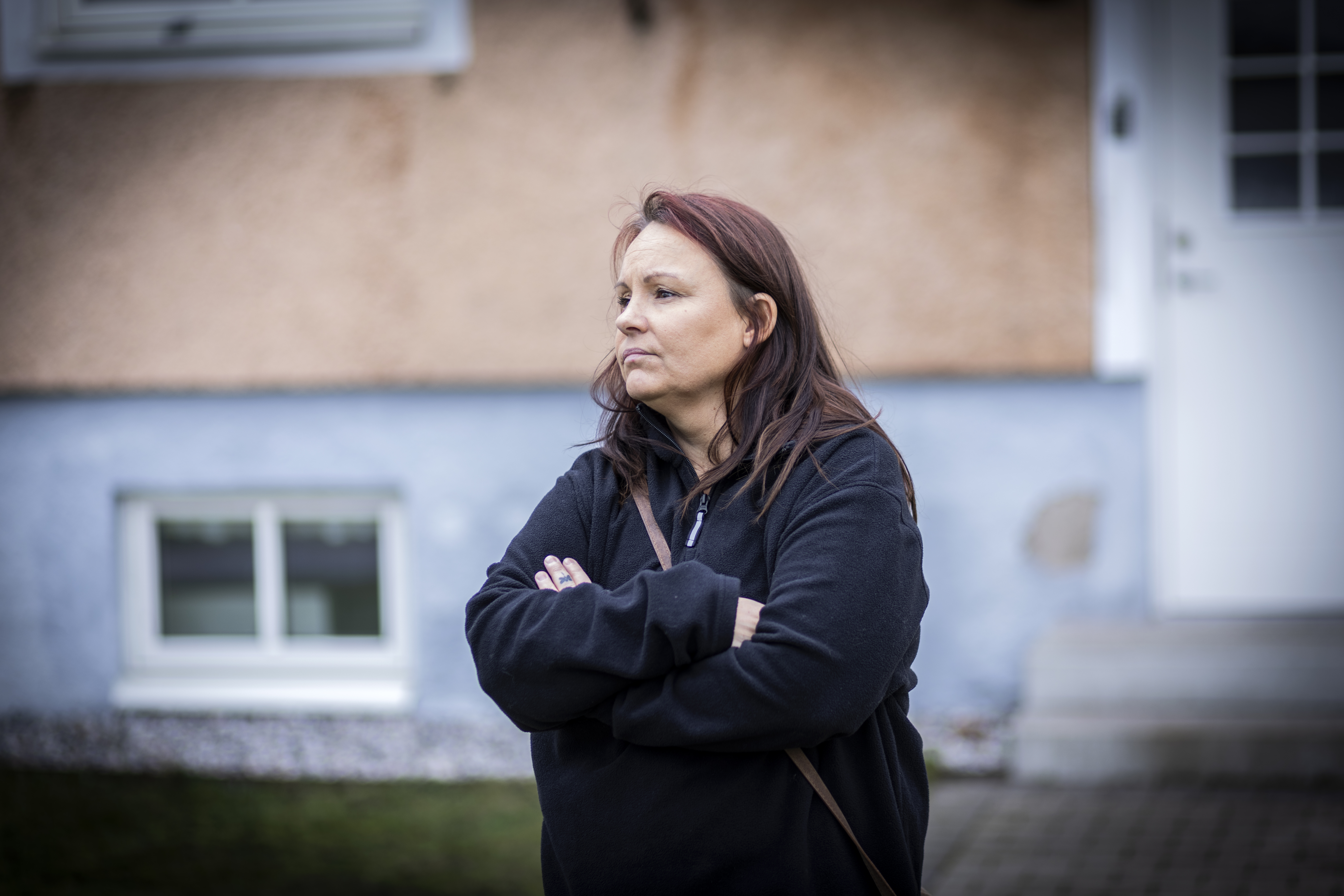 Jessica Axelsson med långt brunt hår står utanför ett hus med armarna i kors och har på sig en svart jacka. Bakom syns fönster, trappa och en vägg med blå kant.