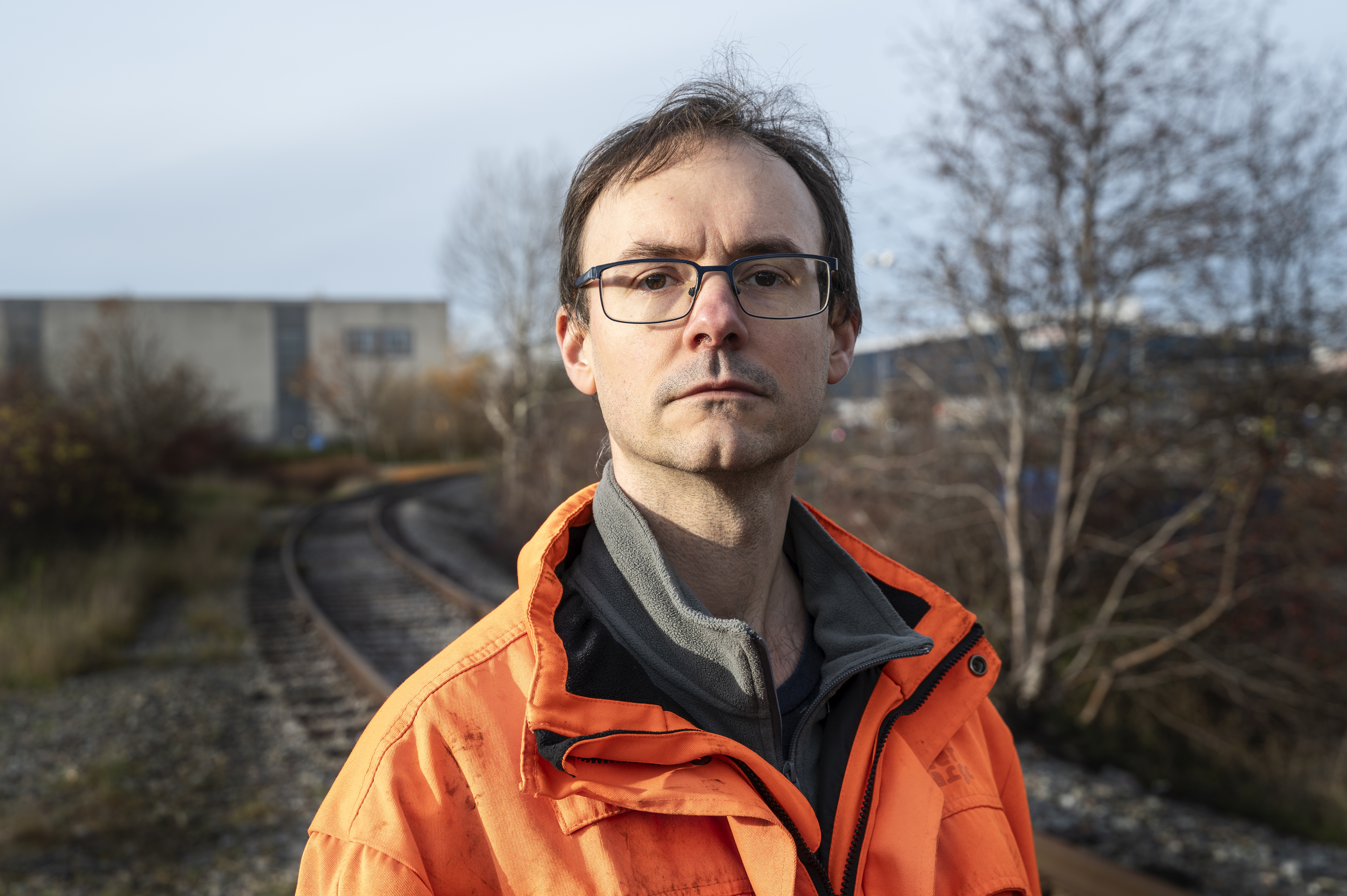 Fredrik Stangel med glasögon och orange jacka står utomhus vid järnvägsspår, med träd utan löv och industribyggnader i bakgrunden.