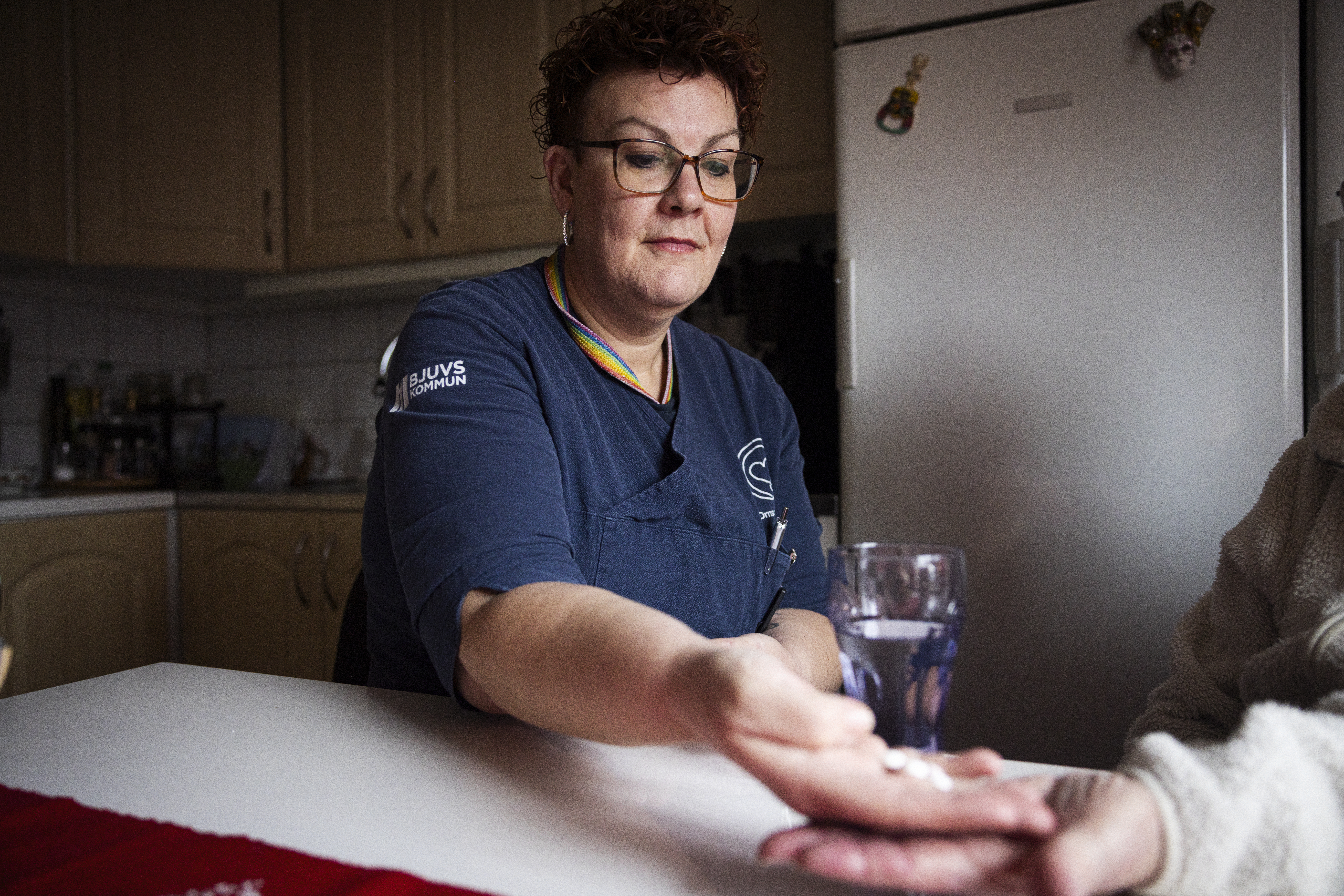 En sjuksköterska i blå arbetskläder räcker fram tabletter till en sittande person i ett kök med ett vattenglas och kylskåp.