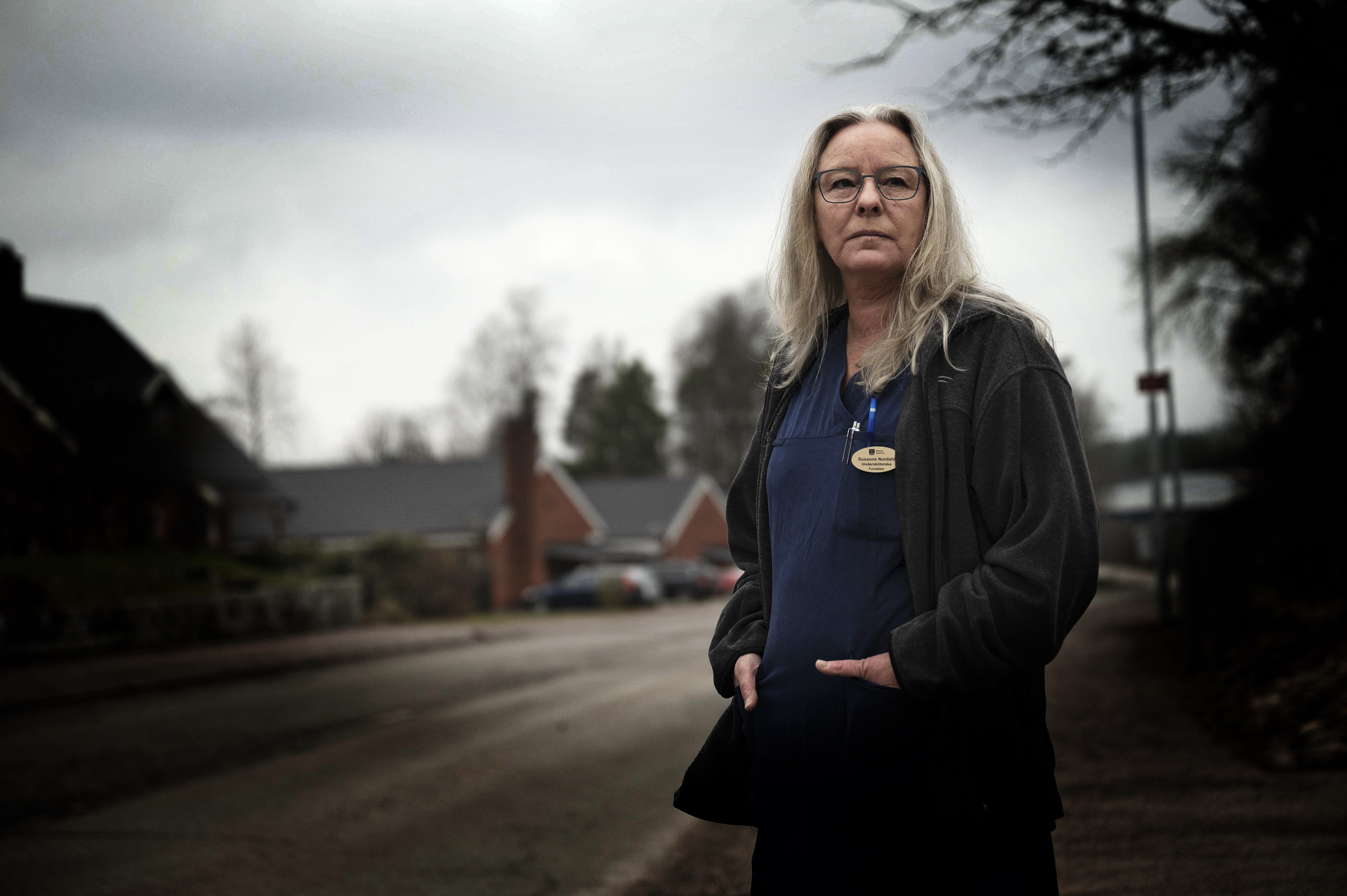 Suzanne Nordahl, med långt grått hår, glasögon och en svart jacka över sina arbetskläder, står på gatan med hus och träd i bakgrunden.