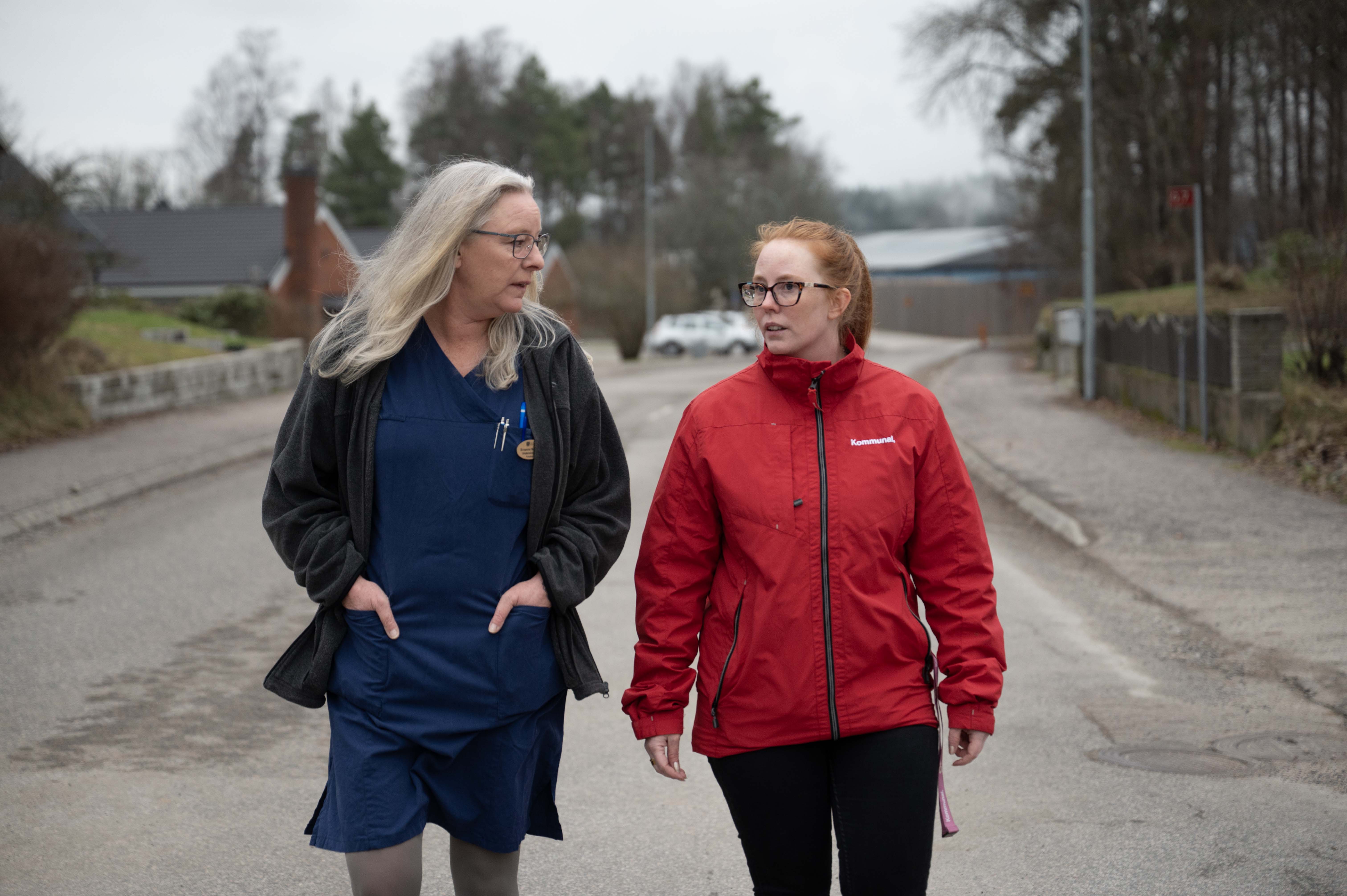 Två kvinnor går och pratar på en gata, den ena i blå arbetskläder och svart jacka, den andra i röd jacka. Träd och hus syns bakom.