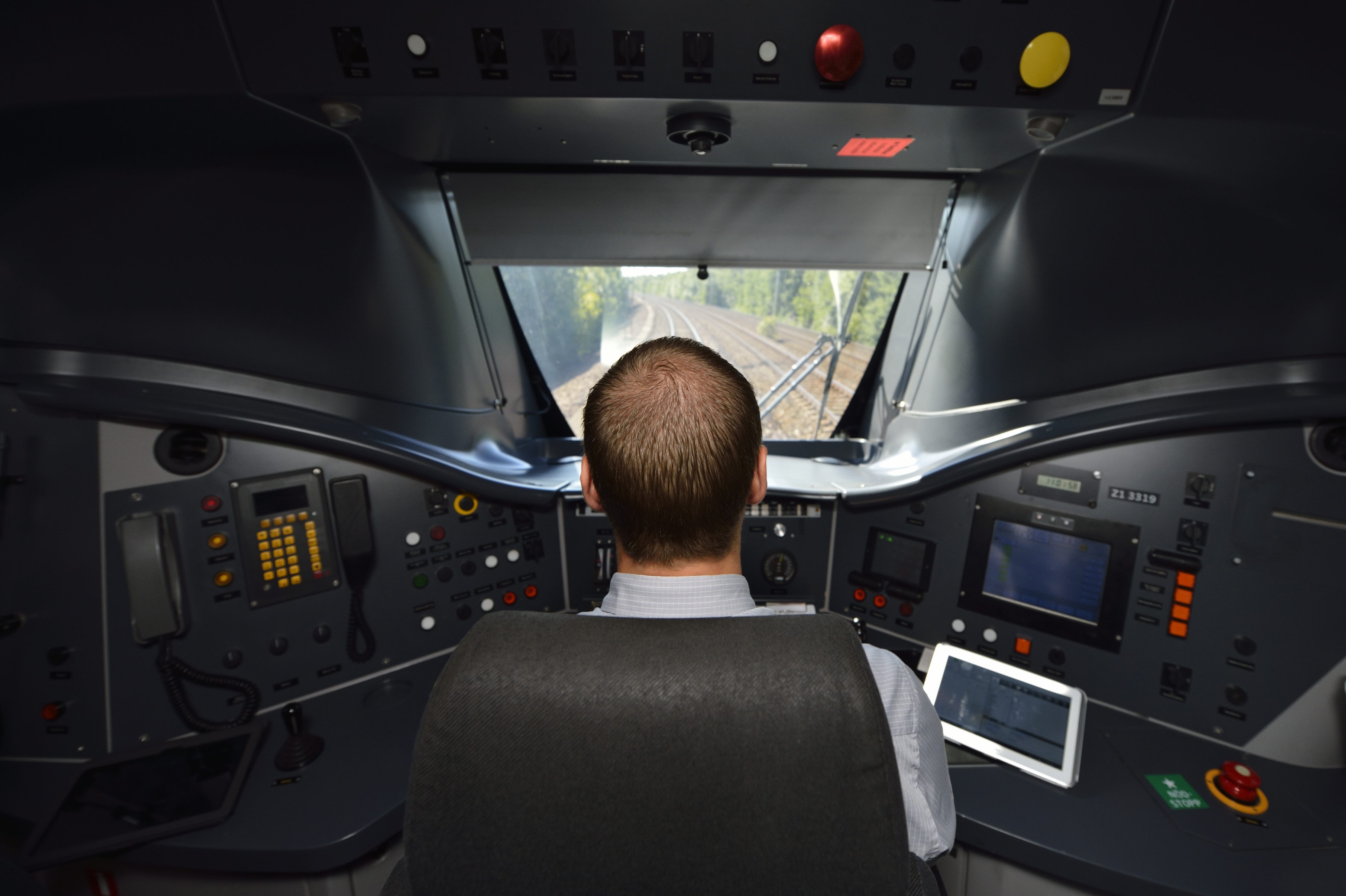 Lokföraren sitter i hytten, omgiven av paneler, skärmar och knappar, vänd framåt. Du kan se järnvägsspåren från fönstret.