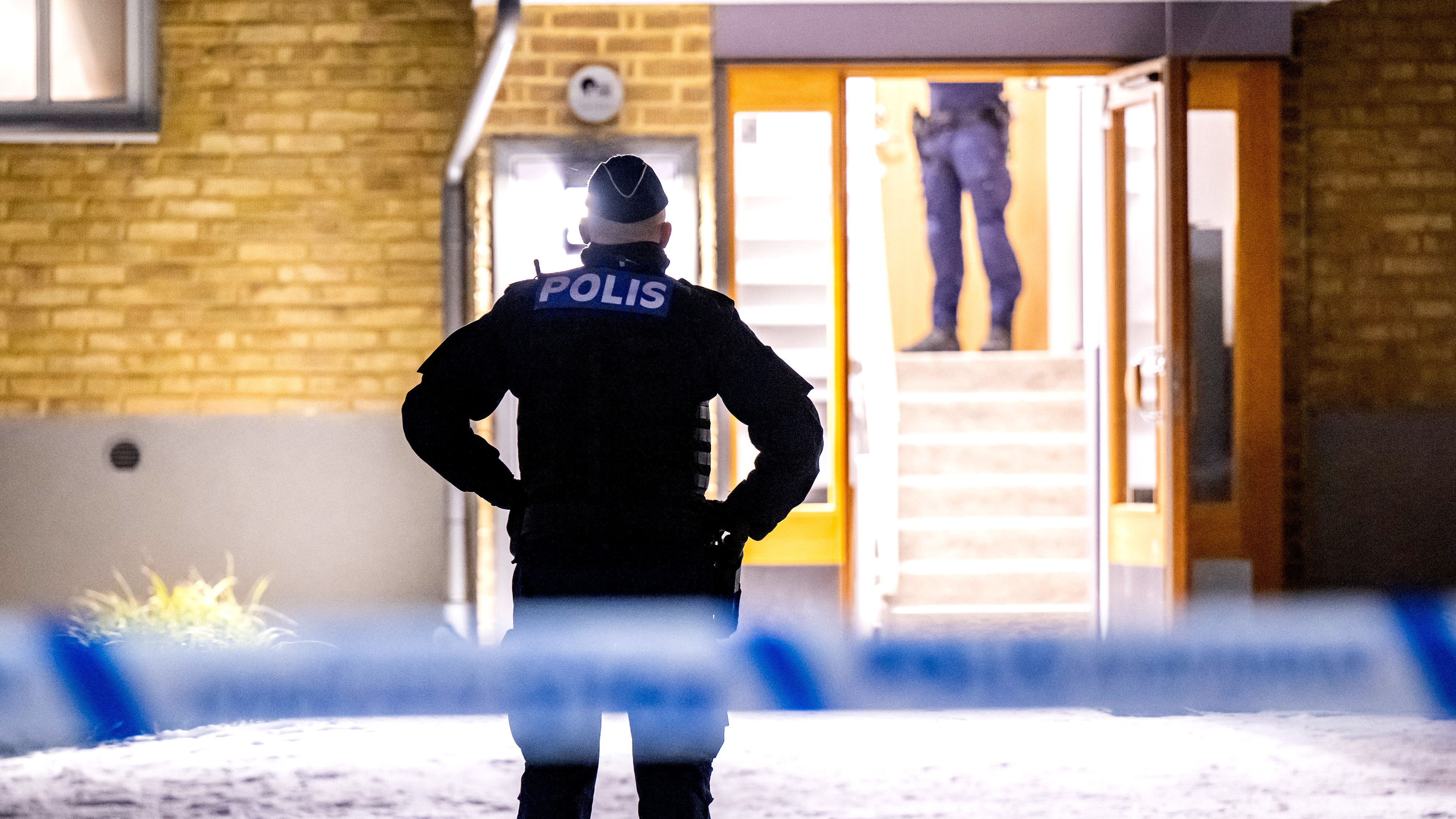 En polis står utanför en byggnad med polistejp i förgrunden och en annan polis syns i ingången.