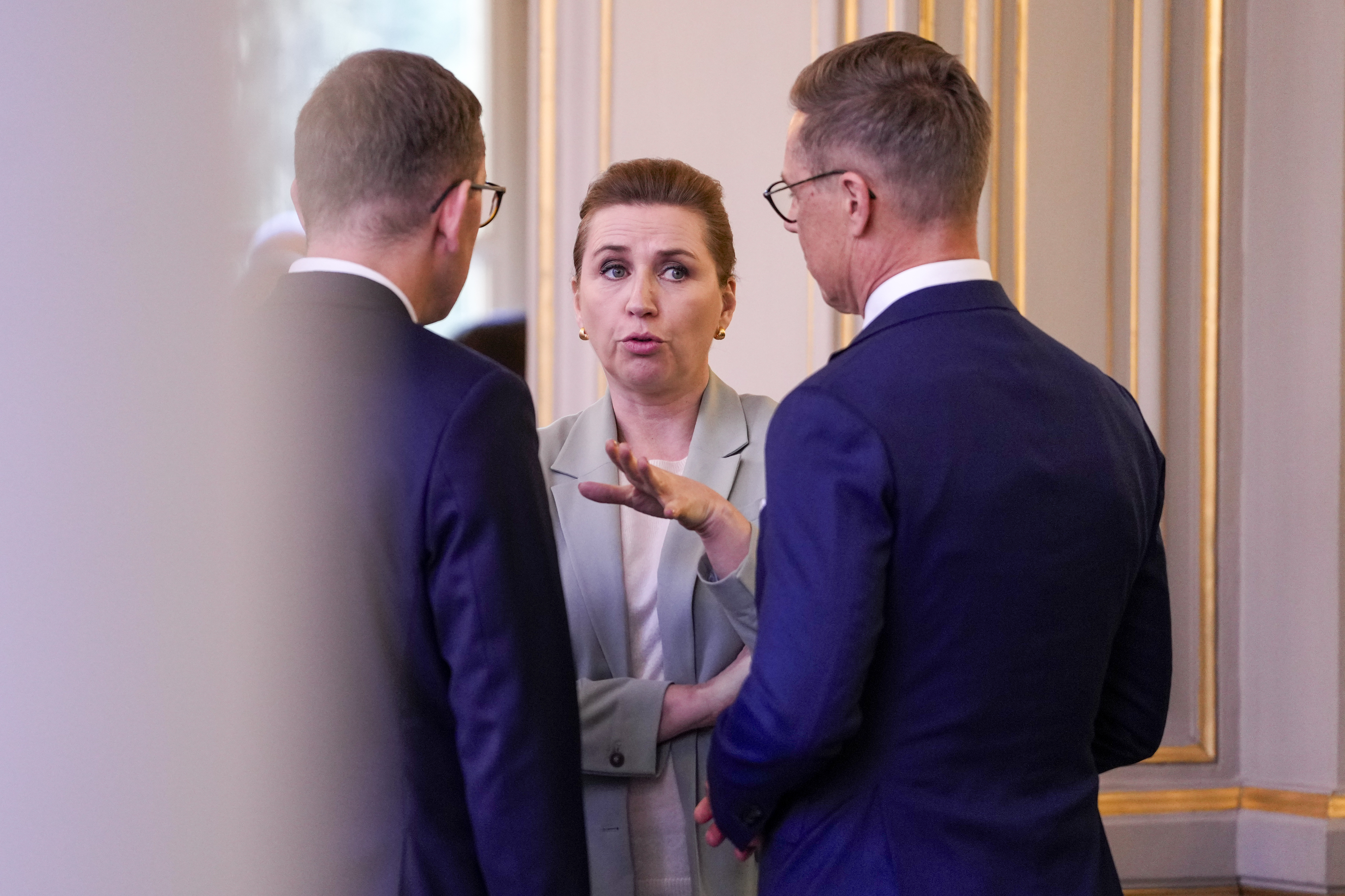 Mette Fredriksen i ljus kostym gestikulerar medan hon talar med två män i mörka kostymer och glasögon i ett rum.