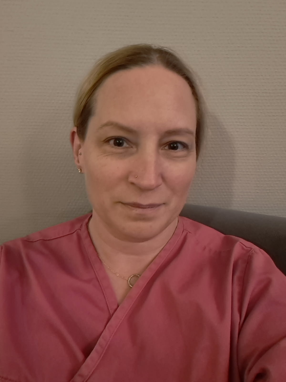 En person med rosa vårdkläder sitter framför en beige vägg och tittar mot kameran med neutral min.