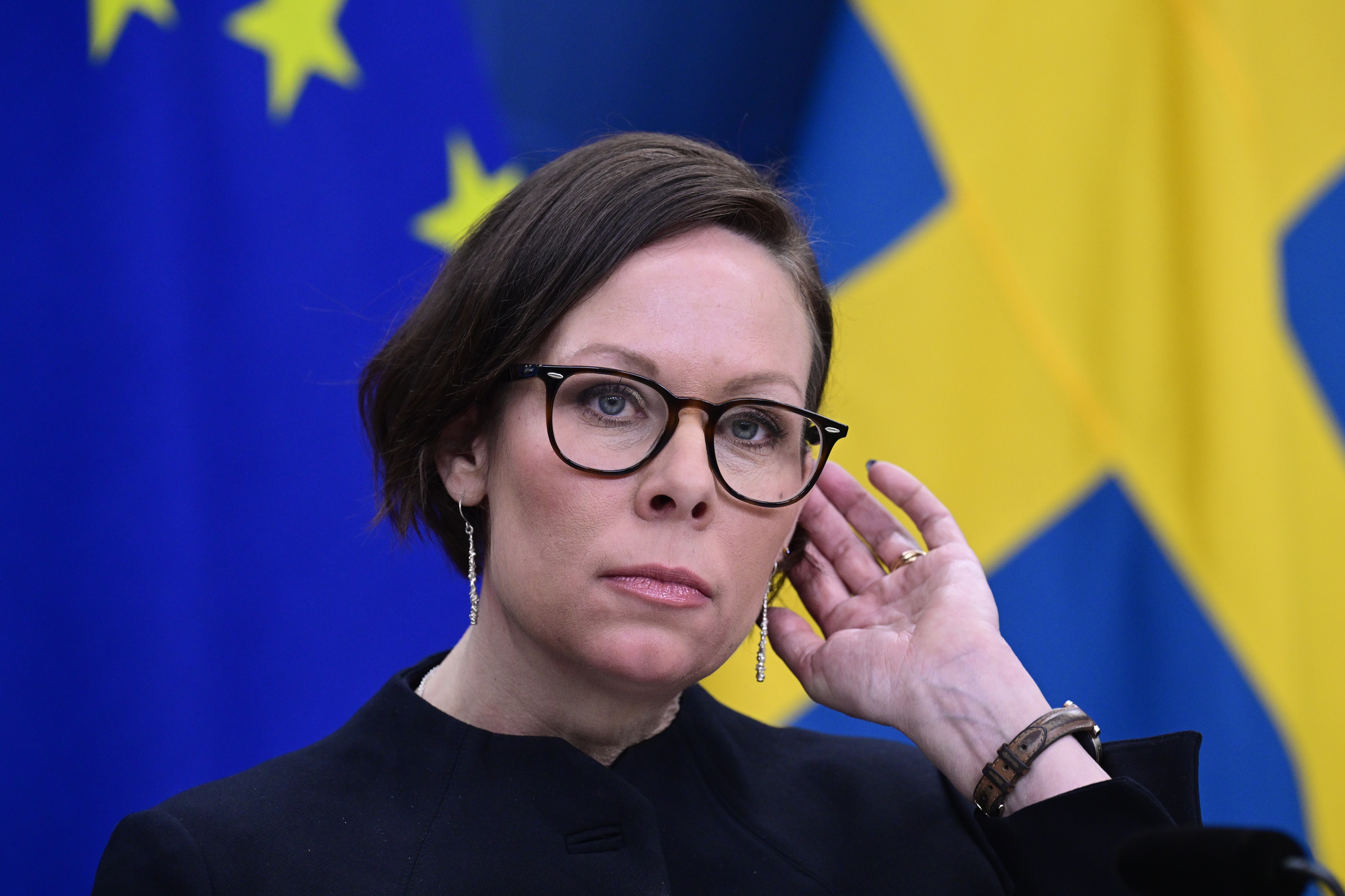 Utrikesminister Maria Malmer Stenergard med glasögon står framför Europeiska unionens och Sveriges flaggor, rör vid sitt öra och tittar framåt.