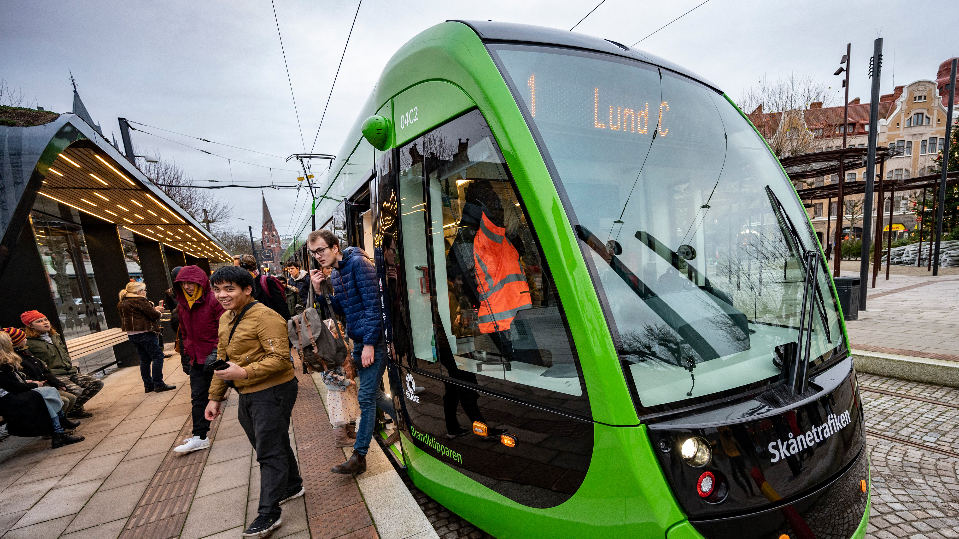 En spårvagn med skylten "Lund C" stannar vid en station. Flera personer går på eller väntar på perrongen i en stadsmiljö.