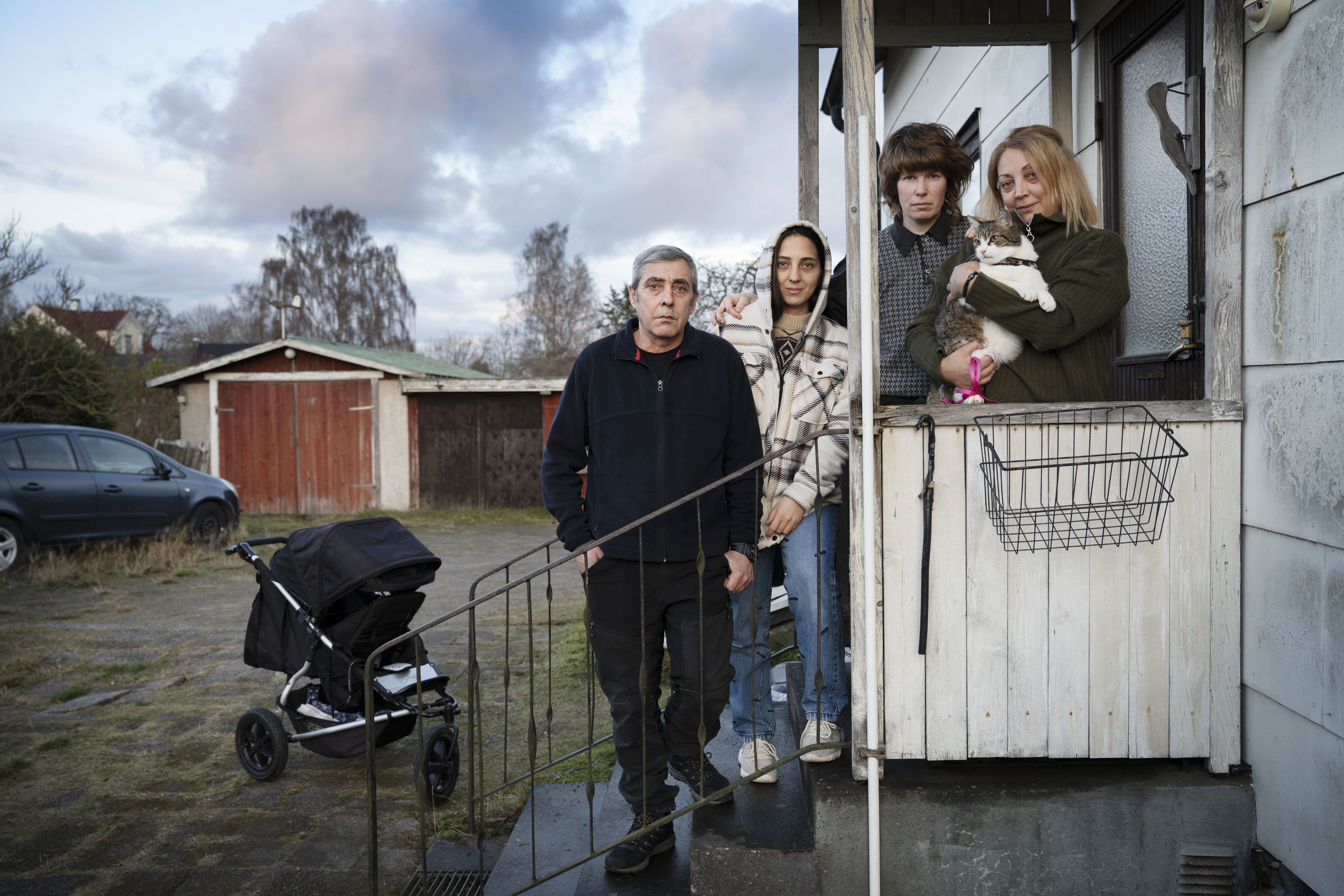 Fyra personer och en katt står på trappan och verandan till ett hus. I bakgrunden syns en barnvagn, bil och skjul.