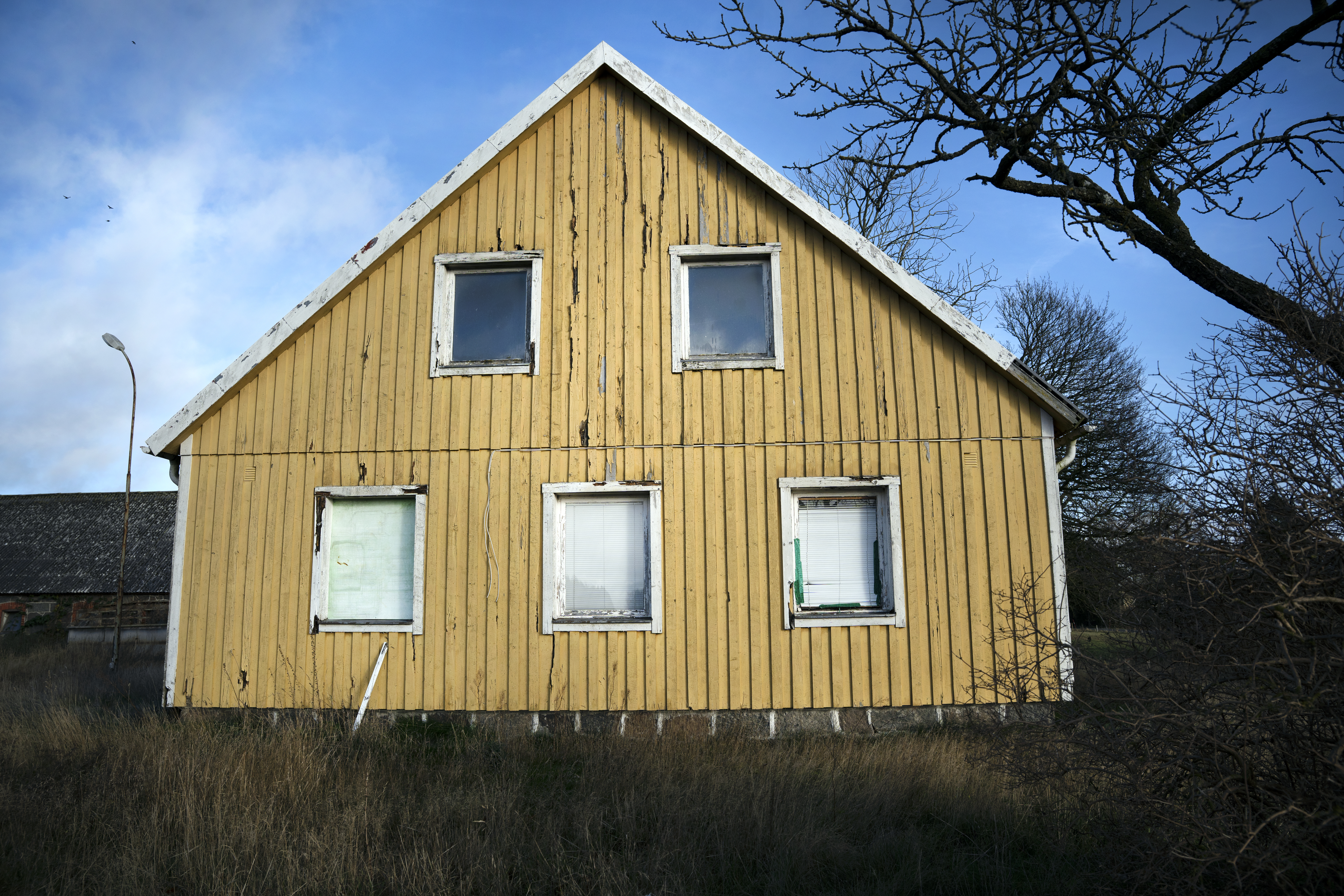 Ett gult slitet trähus med fyra fönster på nedervåningen och två på övervåningen, omgiven av torrt gräs och träd utan löv under blå himmel.
