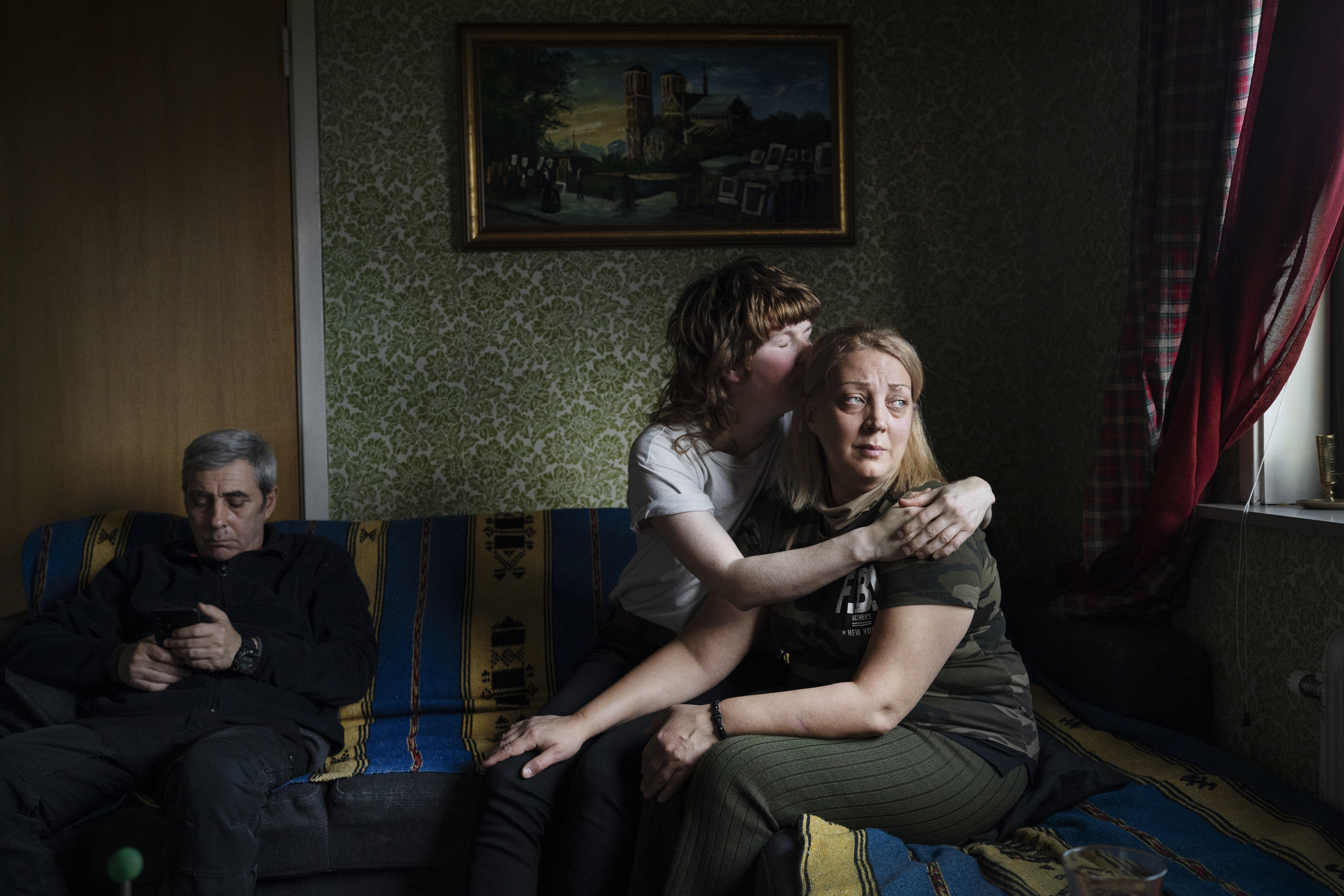 Tre personer sitter i en soffa; Elmer Eklund kramar och viskar till Mariia Koko­sadze, medan Egmont Koko­sadze tittar på sin telefon. En tavla finns på väggen bakom.