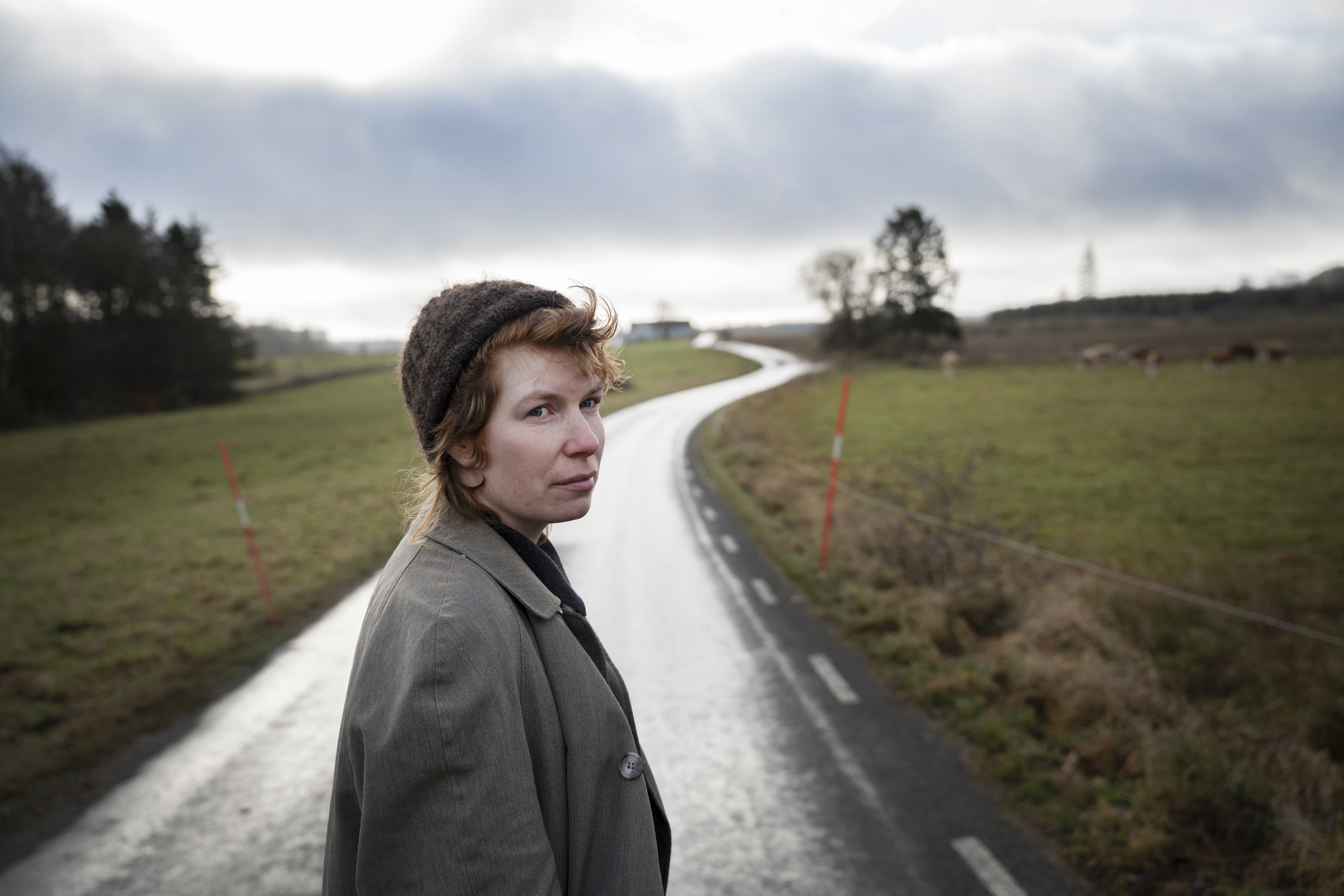 Elmer Eklund med rock och hatt står på en landsväg med åkrar och träd på båda sidor under en molnig himmel.