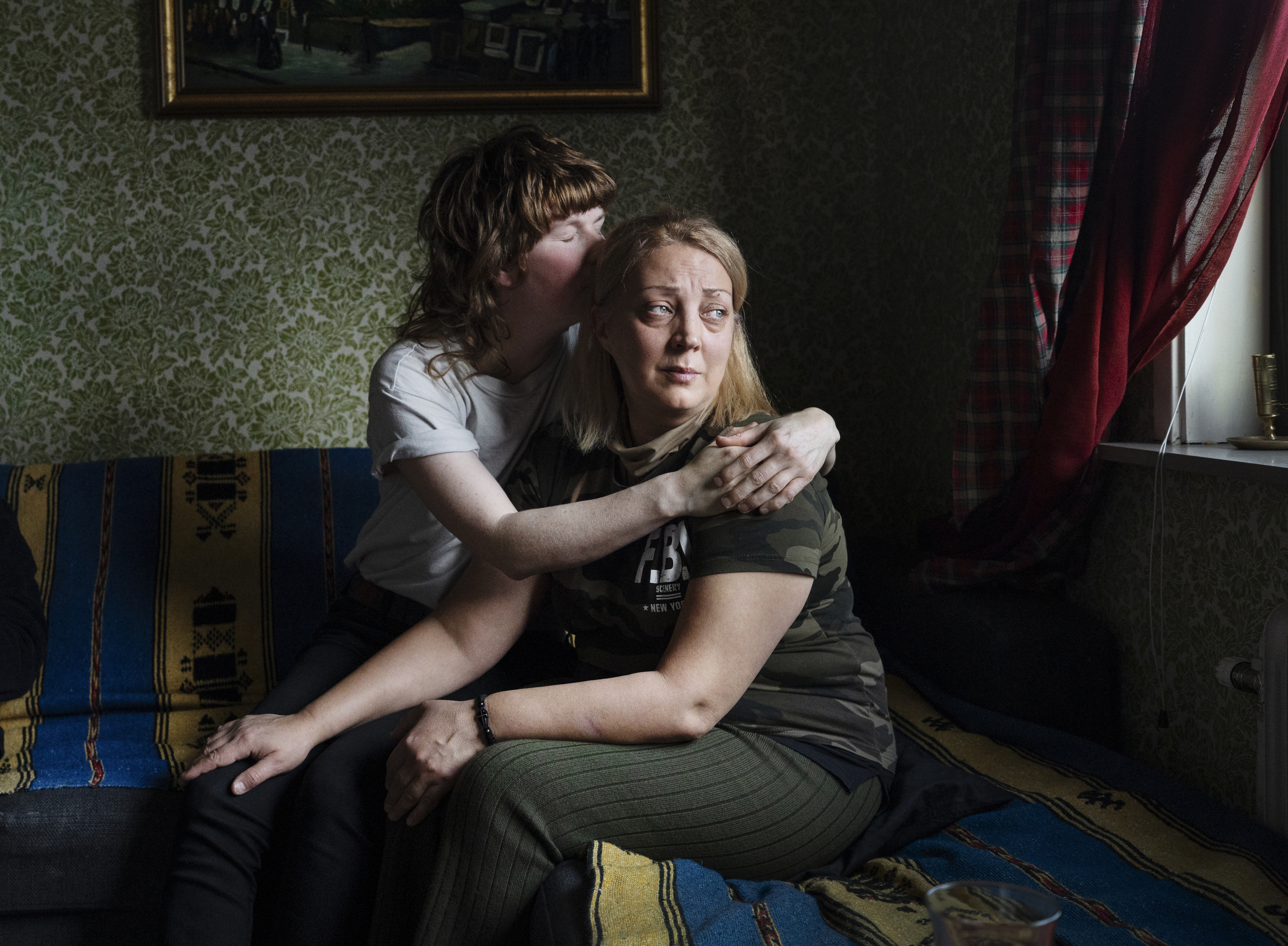 Elmer Eklund kramar och tröstar Mariia Kokosadze som sitter på en soffa i ett rum med mönstrad tapet och ett fönster med röda gardiner.