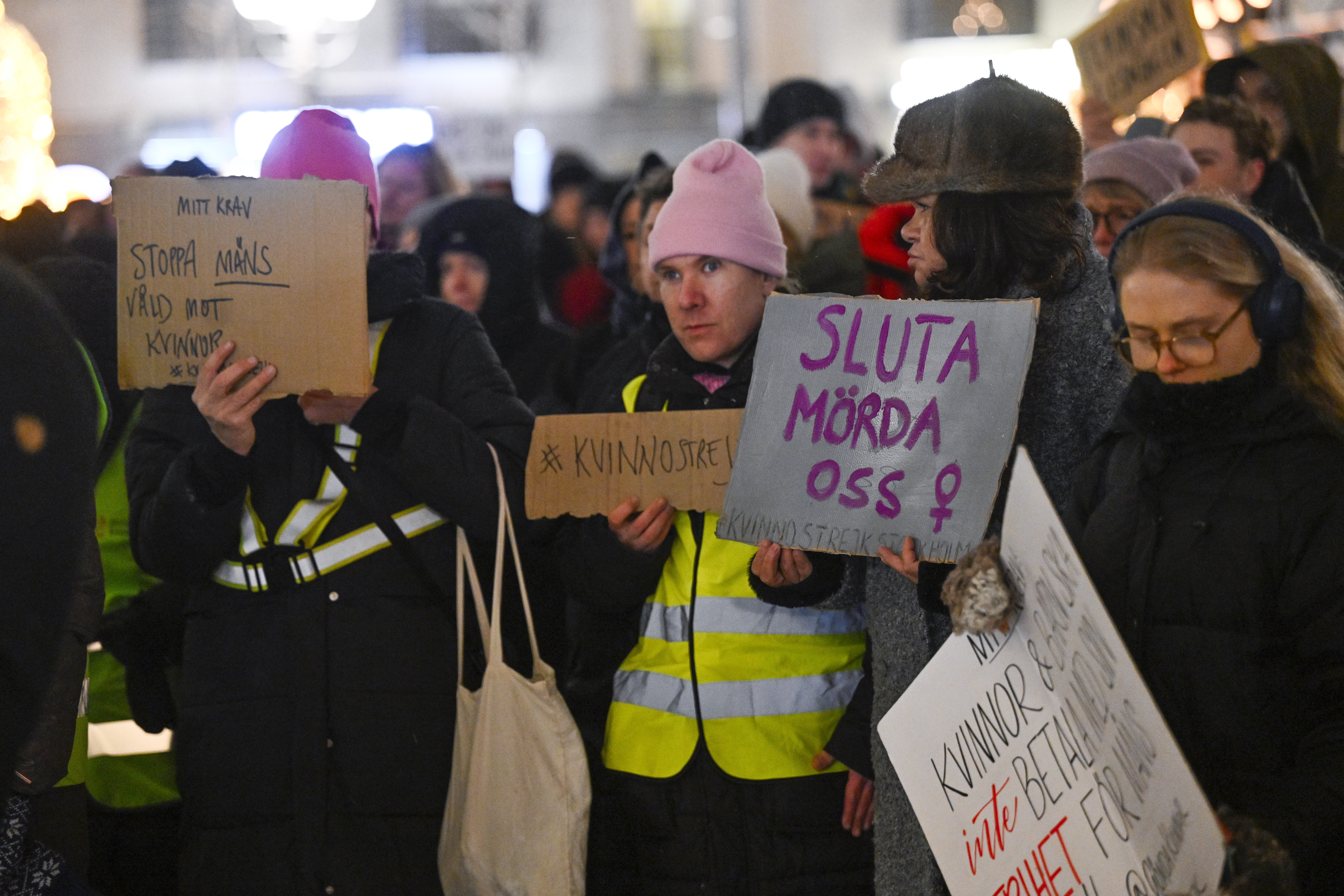 En grupp människor på en protest håller skyltar med svenska budskap som "Stop killing us" och "#WomensStrike". Några har västar och rosa mössor.