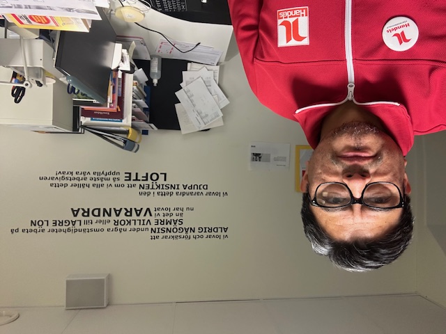 Man med röd jacka och glasögon står i ett kontor med skrivbord, papper och svensk text på väggen bakom sig.