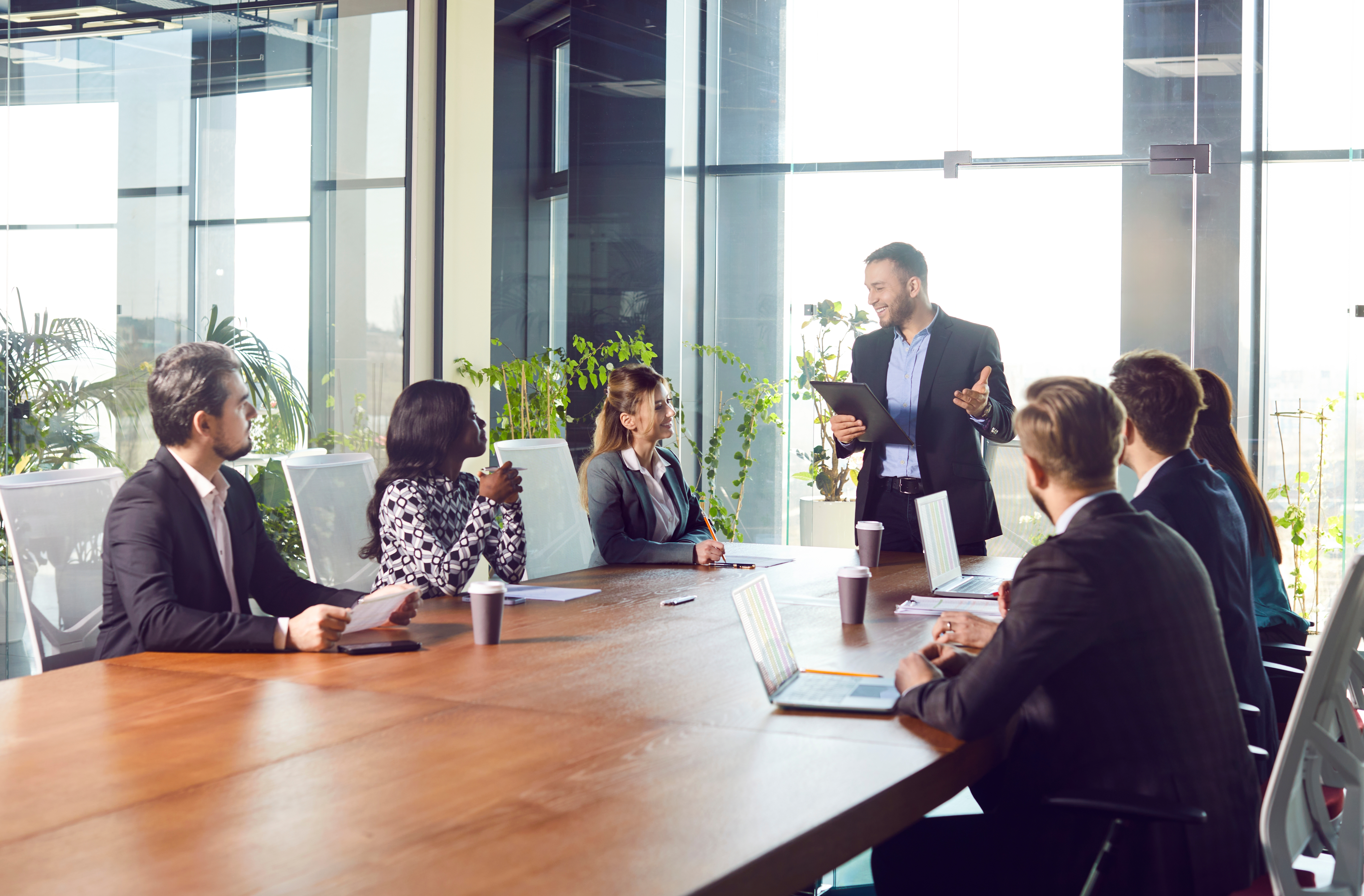 Sex personer sitter runt ett konferensbord medan en man står och talar med en surfplatta. På bordet finns laptops, papper och kaffekoppar.