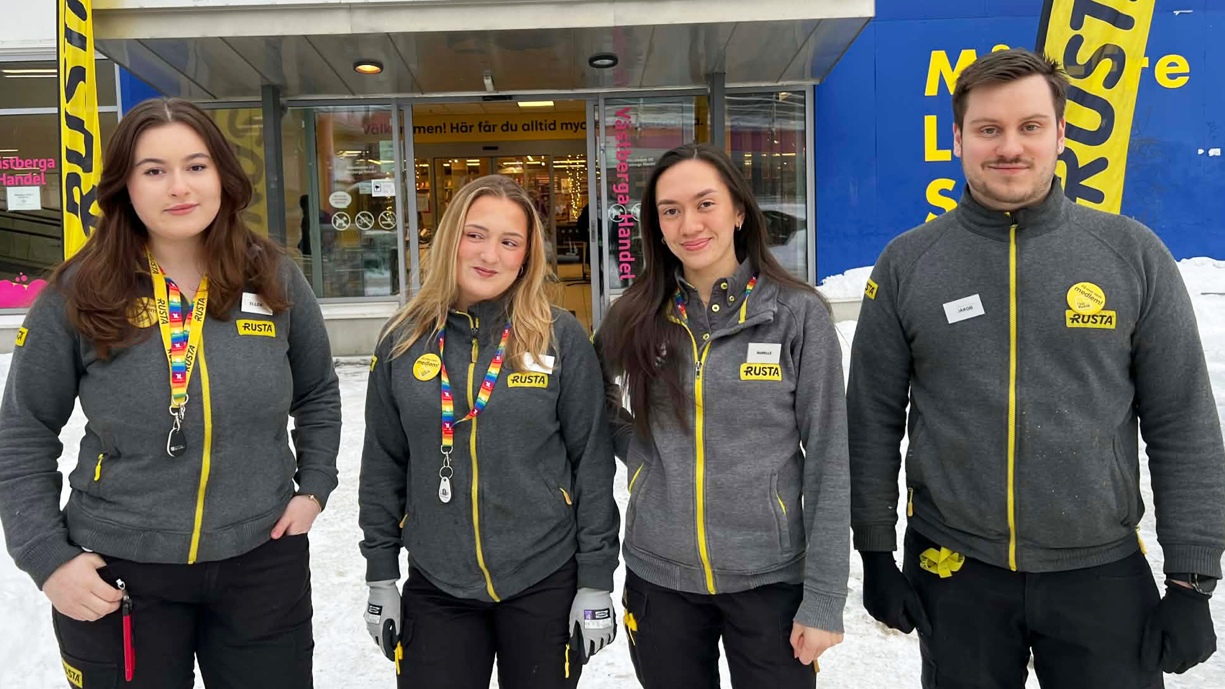 Fyra anställda i grå och gula Rusta-uniformer står utanför en byggnad i snön och tittar mot kameran.