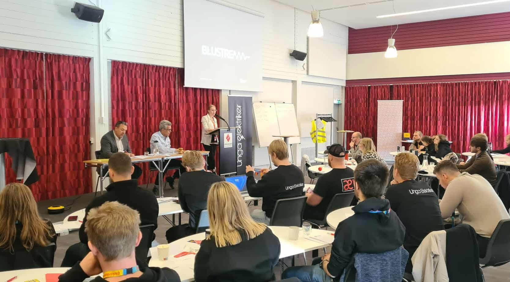 En talare står vid ett podium framför sittande deltagare i en konferenssal, med två paneldeltagare och en skärm bakom.
