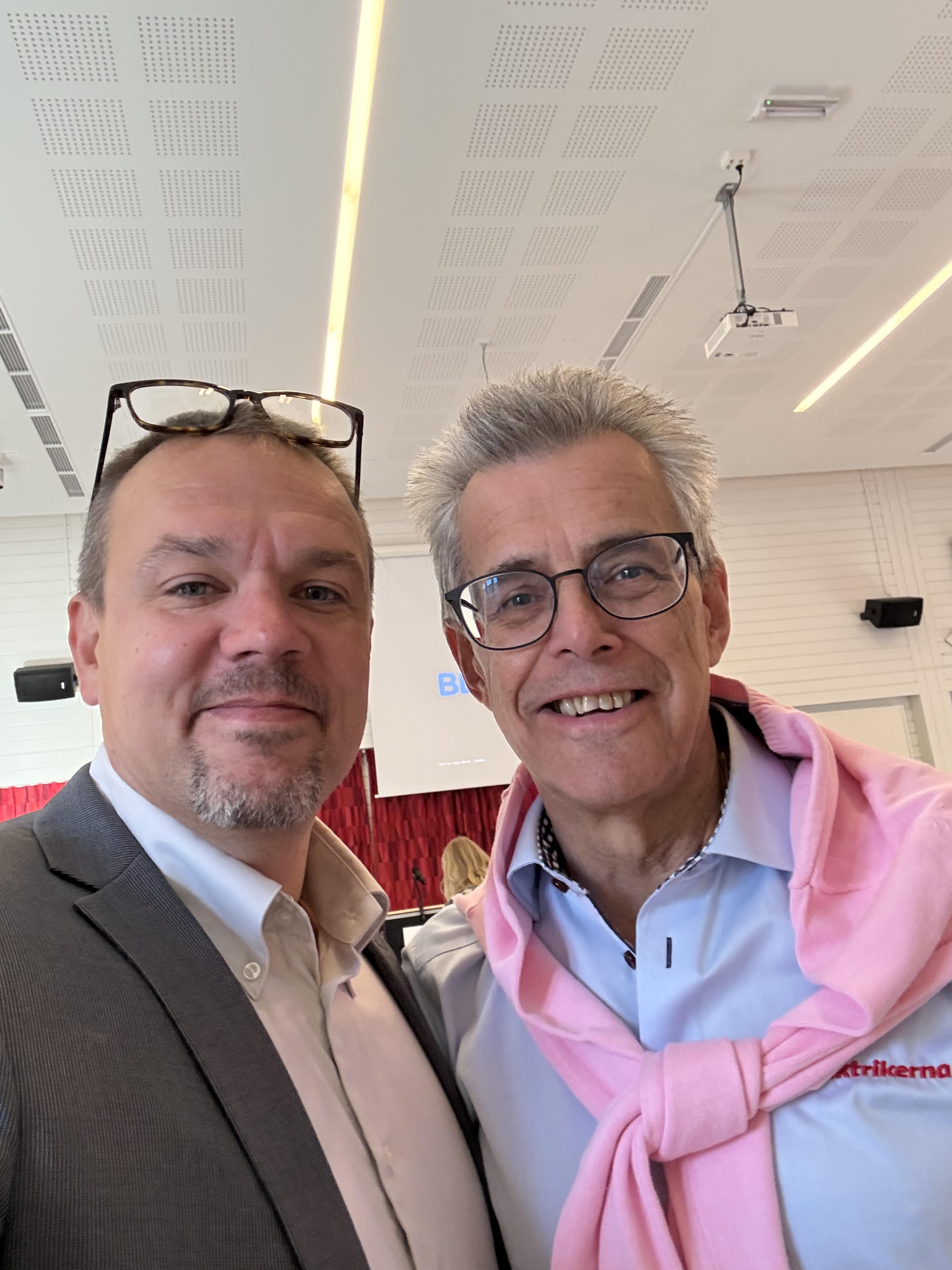 Två män poserar tillsammans inomhus; en har kostym och glasögon, den andra har glasögon och en rosa tröja över axlarna.