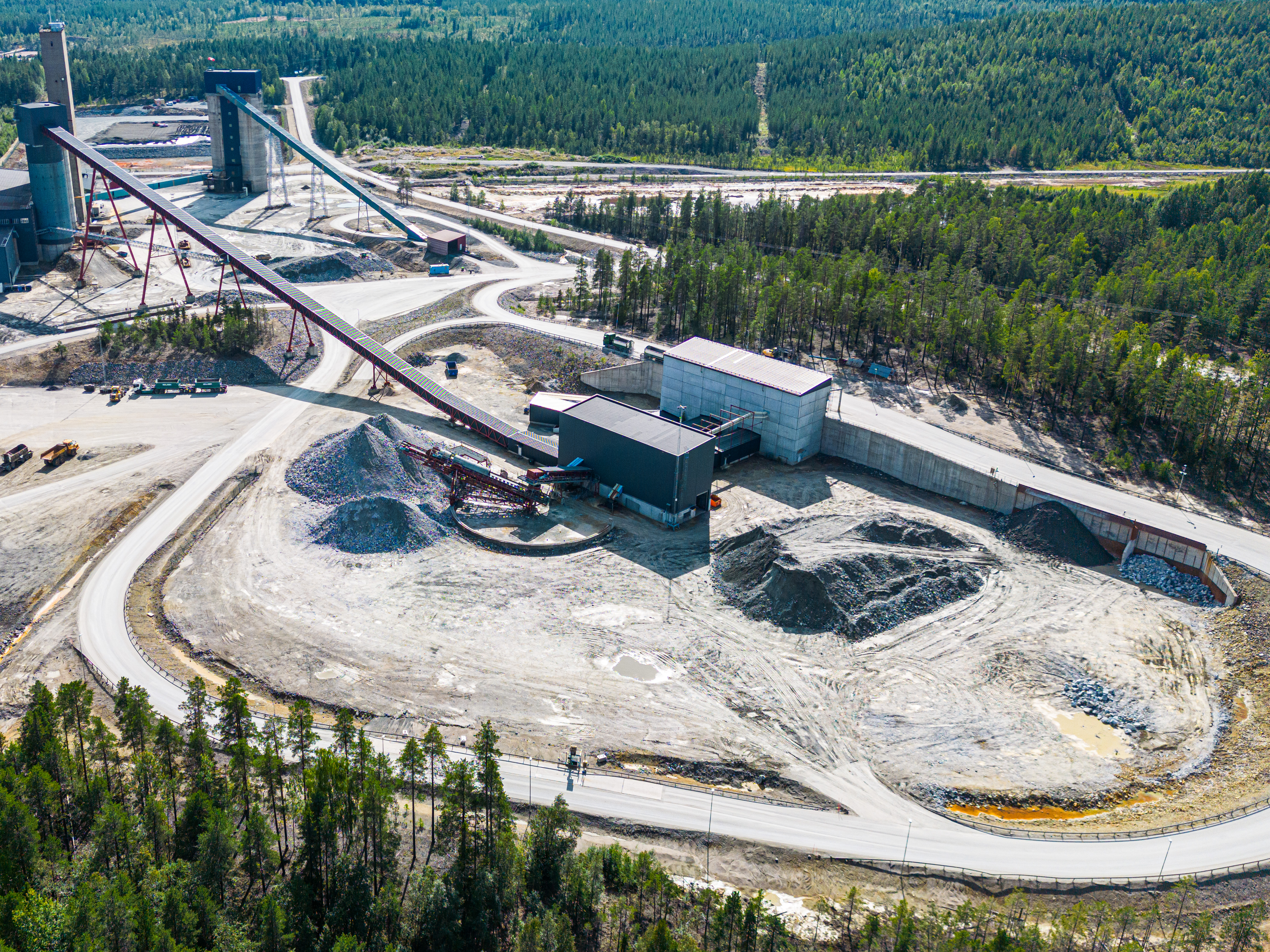 Flygbild av en industriläggning med transportband, lagerbyggnader och högar av råmaterial, omgiven av skog.
