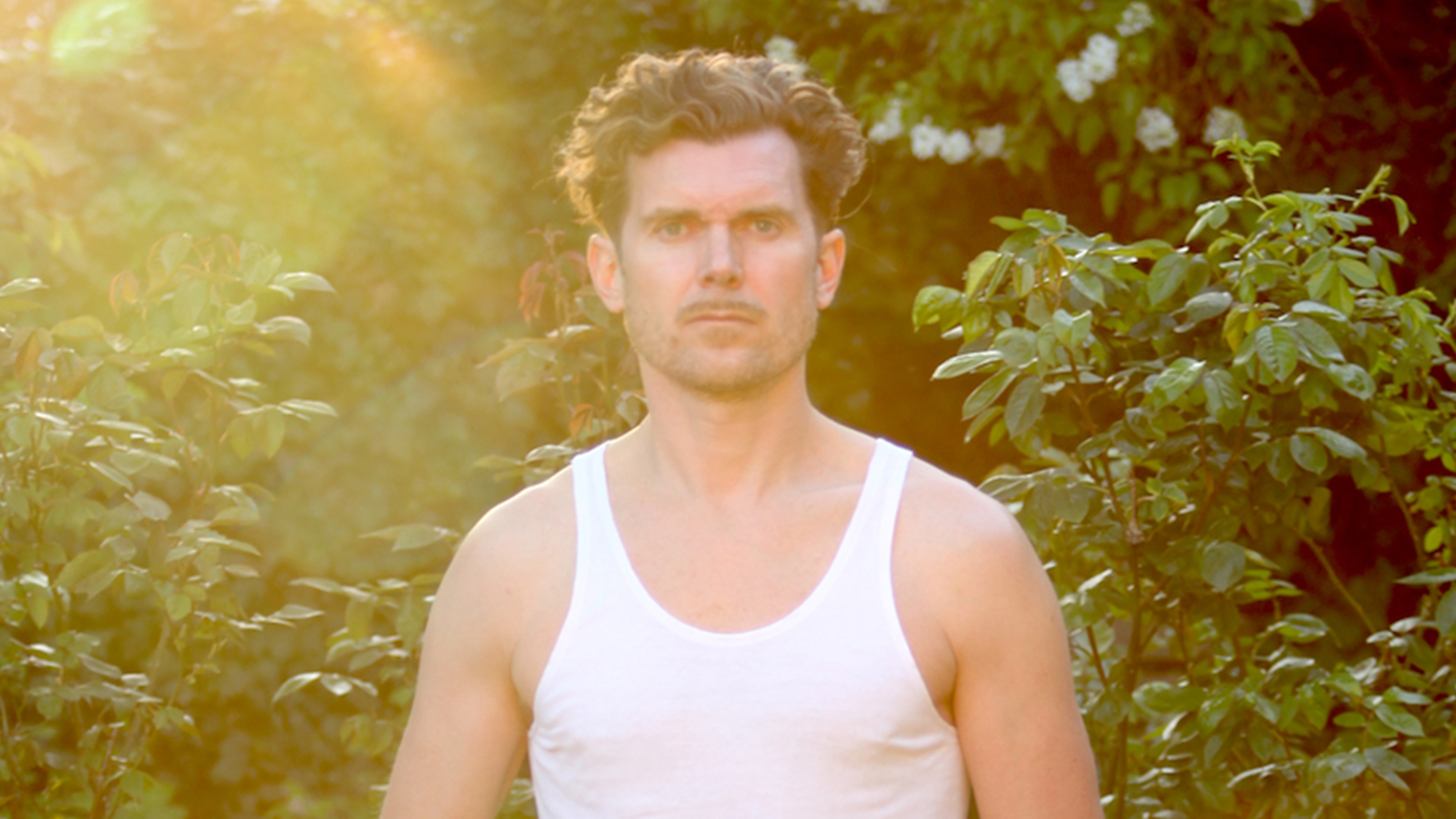 En man i vitt linne står utomhus framför gröna buskar med solljus från vänster sida av bilden.