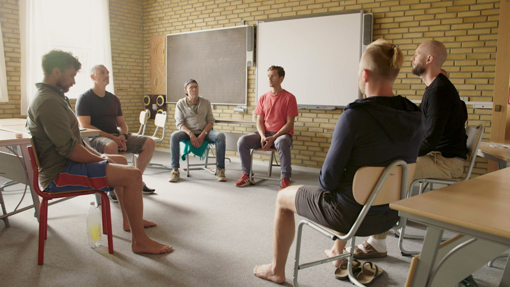 Sex män sitter i en cirkel i ett klassrum med tegelväggar, skrivbord och whiteboards och deltar i en gruppdiskussion.
