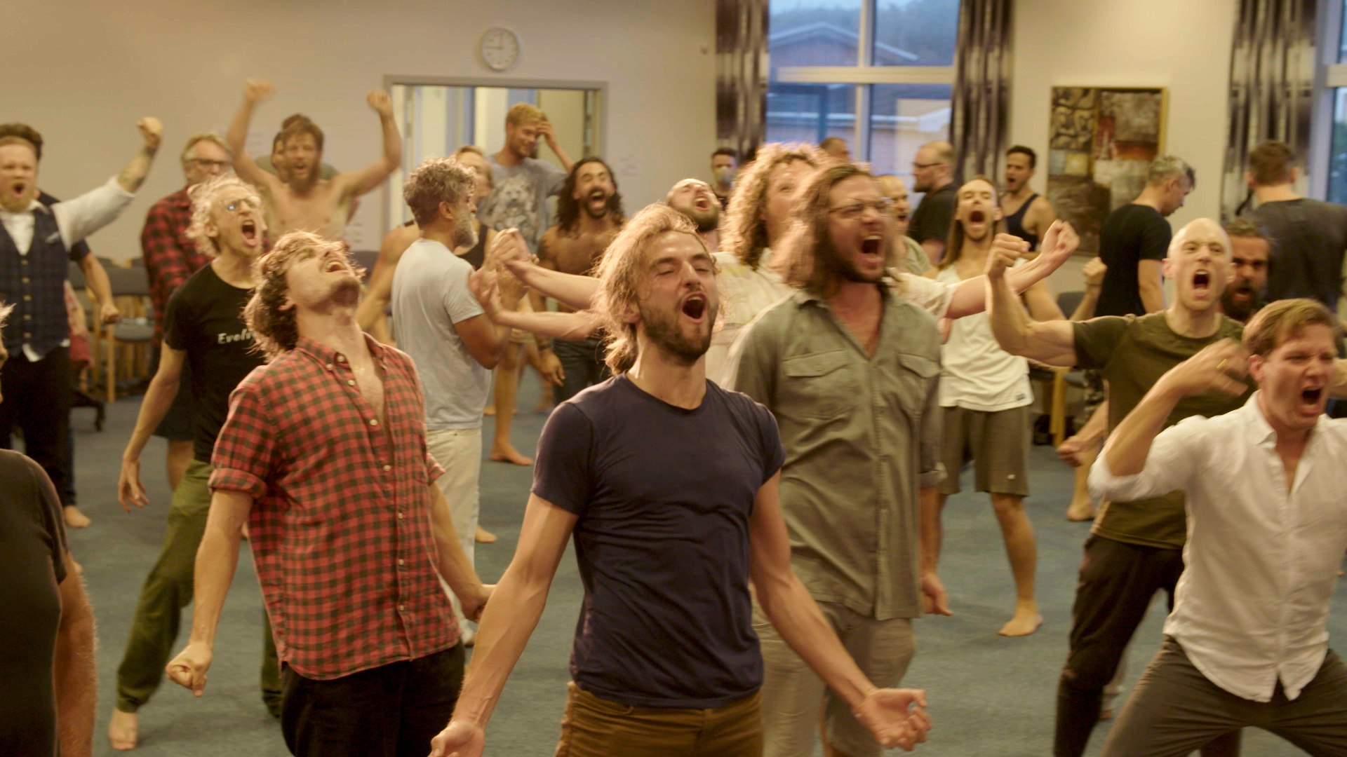 En grupp män i vardagskläder står i ett rum och ropar eller sjunger tillsammans med gester.
