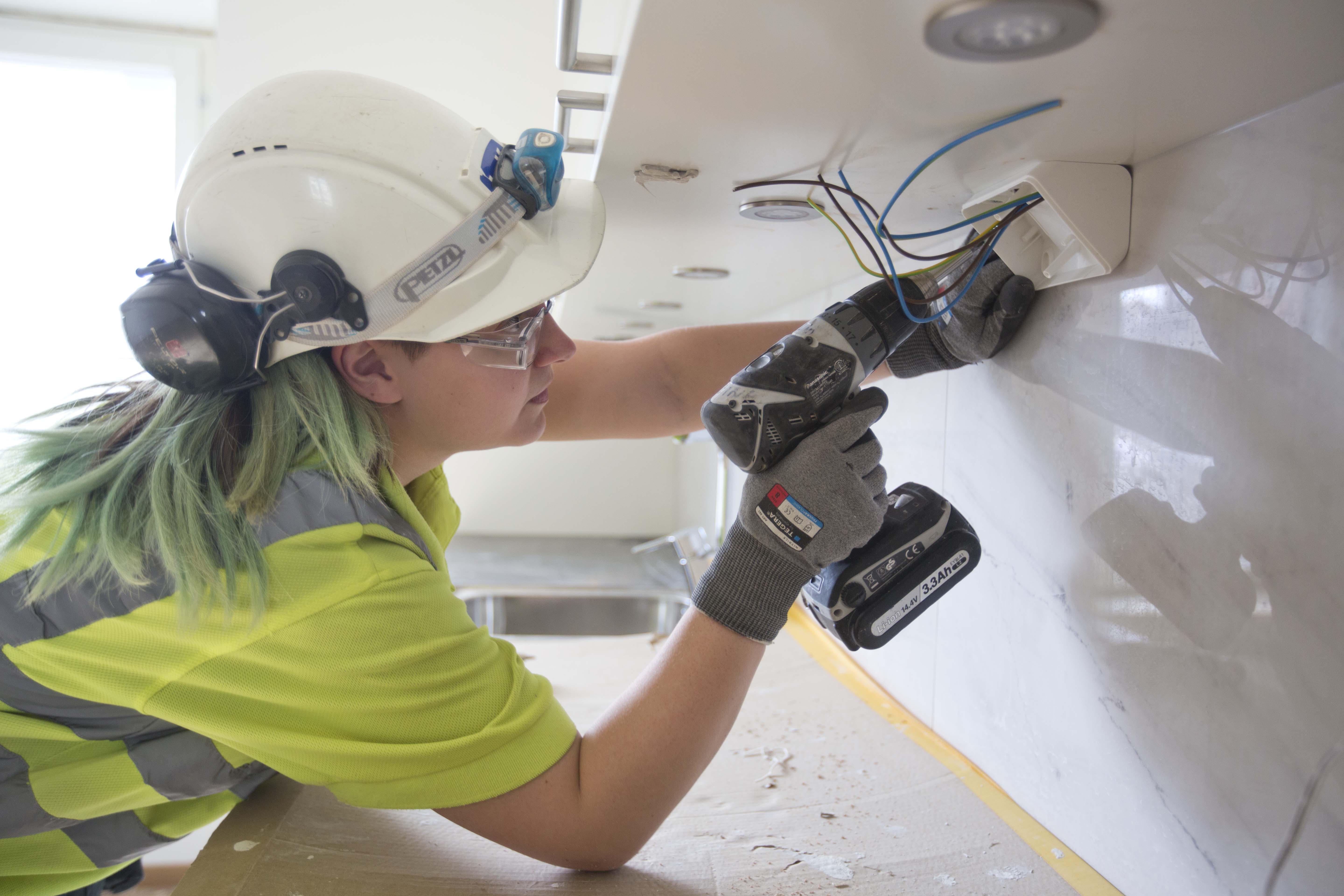 En byggnadsarbetare som bär skyddsutrustning använder en borr och hanterar elkablar under en arbetsbänk.