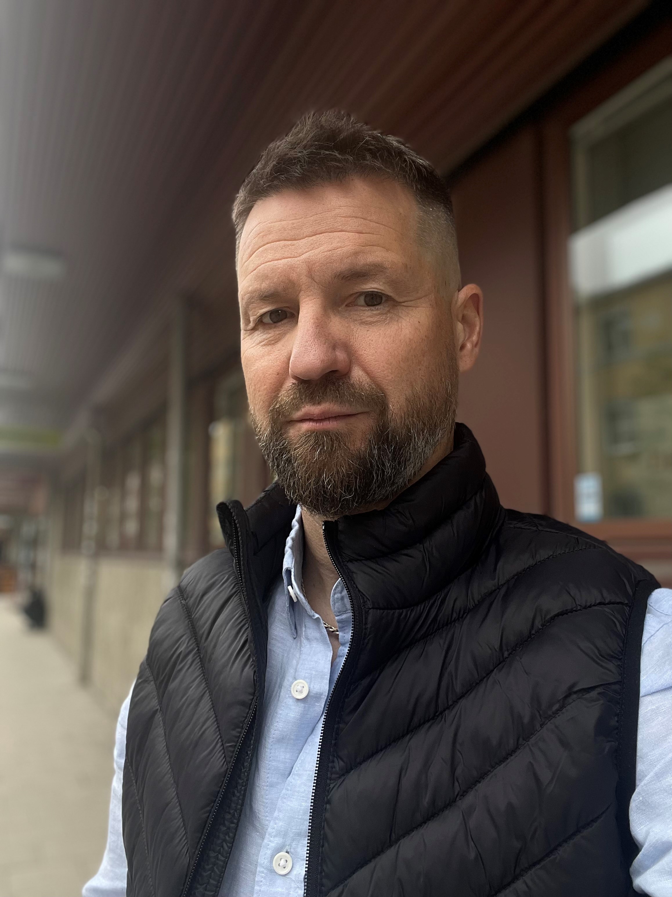 En man med kort hår och skägg, iförd ljusblå skjorta och svart väst, står utomhus vid en byggnad och tittar mot kameran.