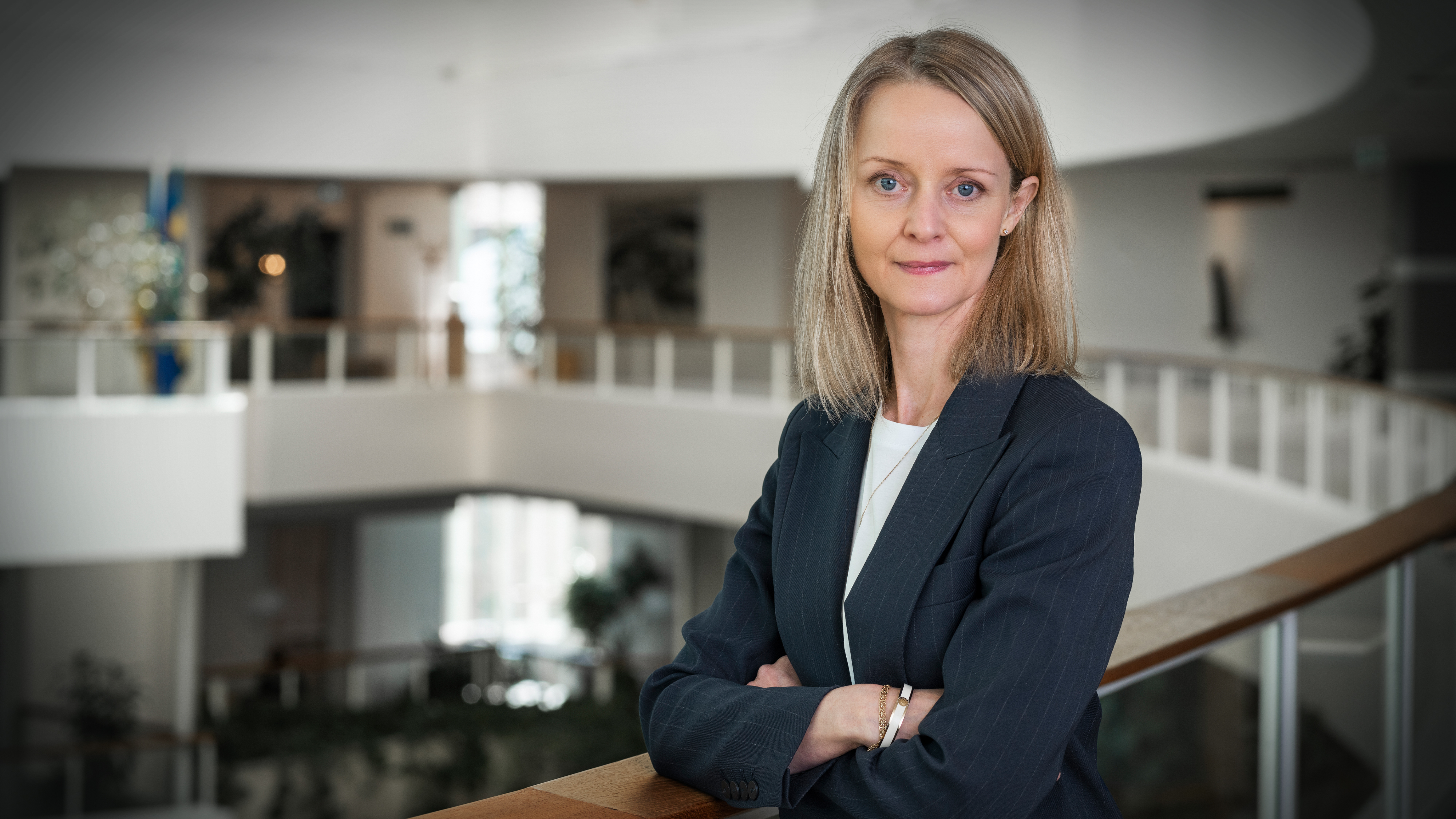 En kvinna i mörk kostym står med armarna i kors och lutar sig mot ett räcke inomhus.