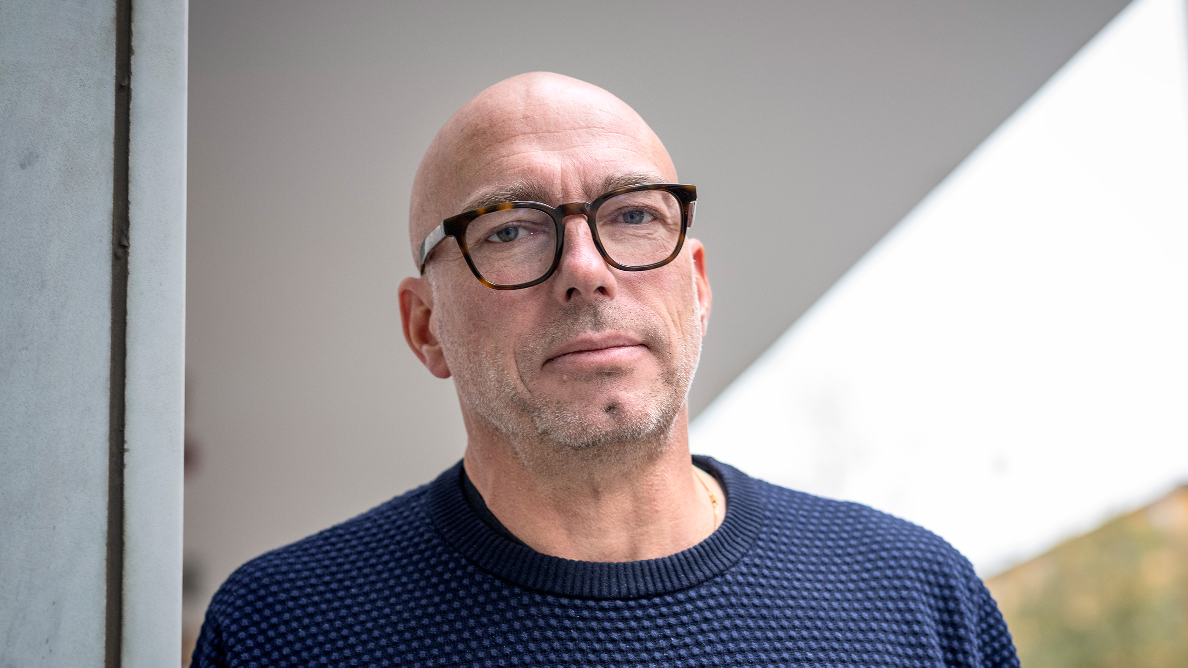 En man som bär glasögon och en blå skjorta står nära en byggnad utomhus och tittar på kameran. Stadsbakgrund med byggnader och skyltar.