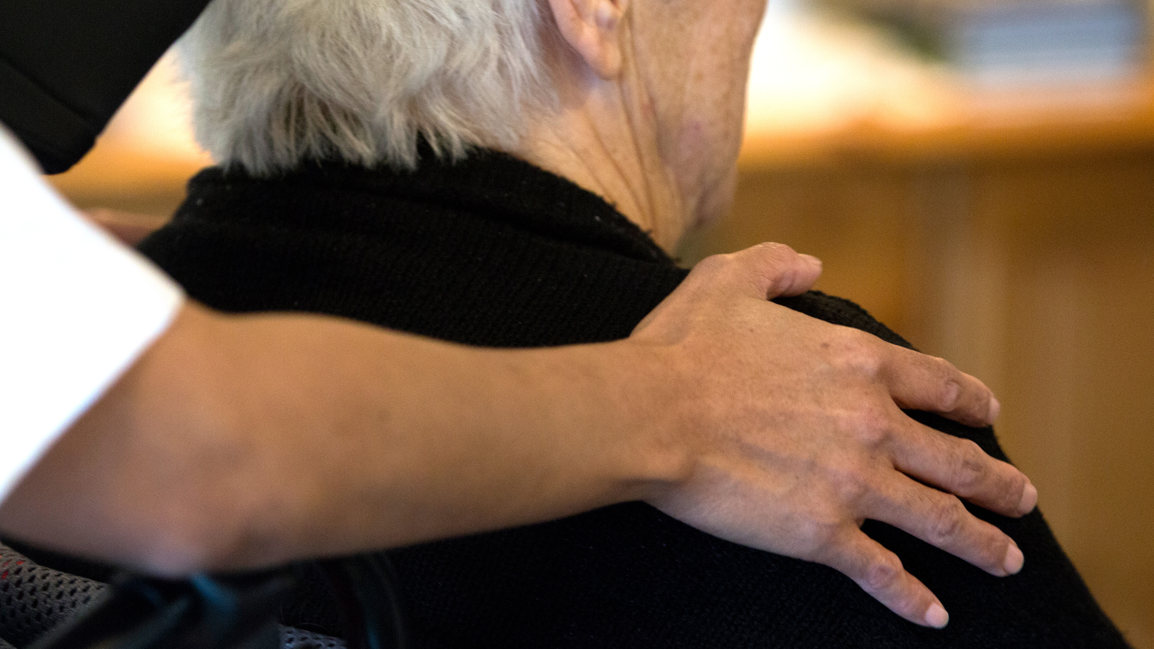 En person lägger sin hand på axeln av en äldre vuxen med grått hår inomhus, vilket visar stöd.