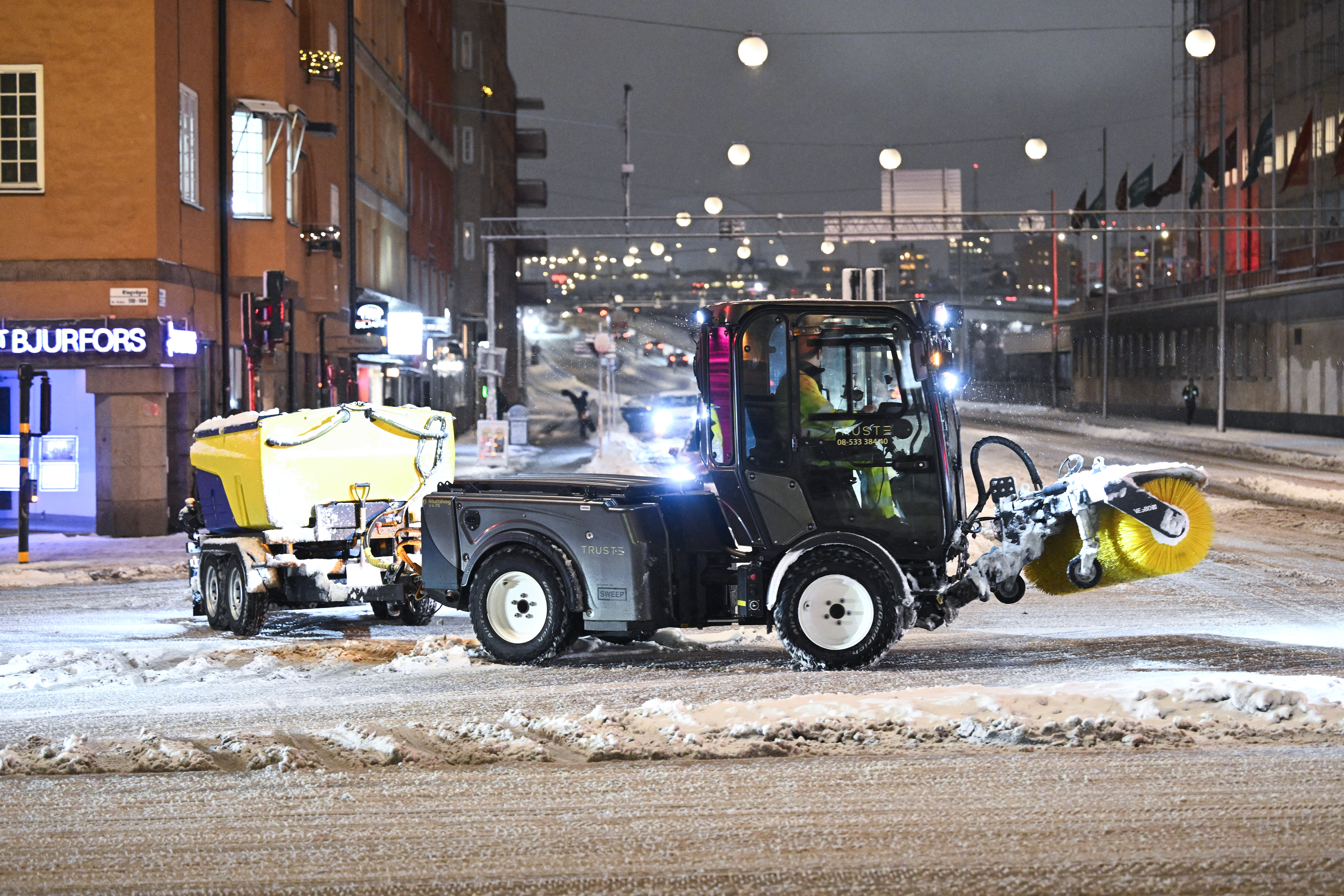 En snöplog med roterande borste tar bort snö från en stadsgata på natten, gatlyktor och byggnader syns i bakgrunden.