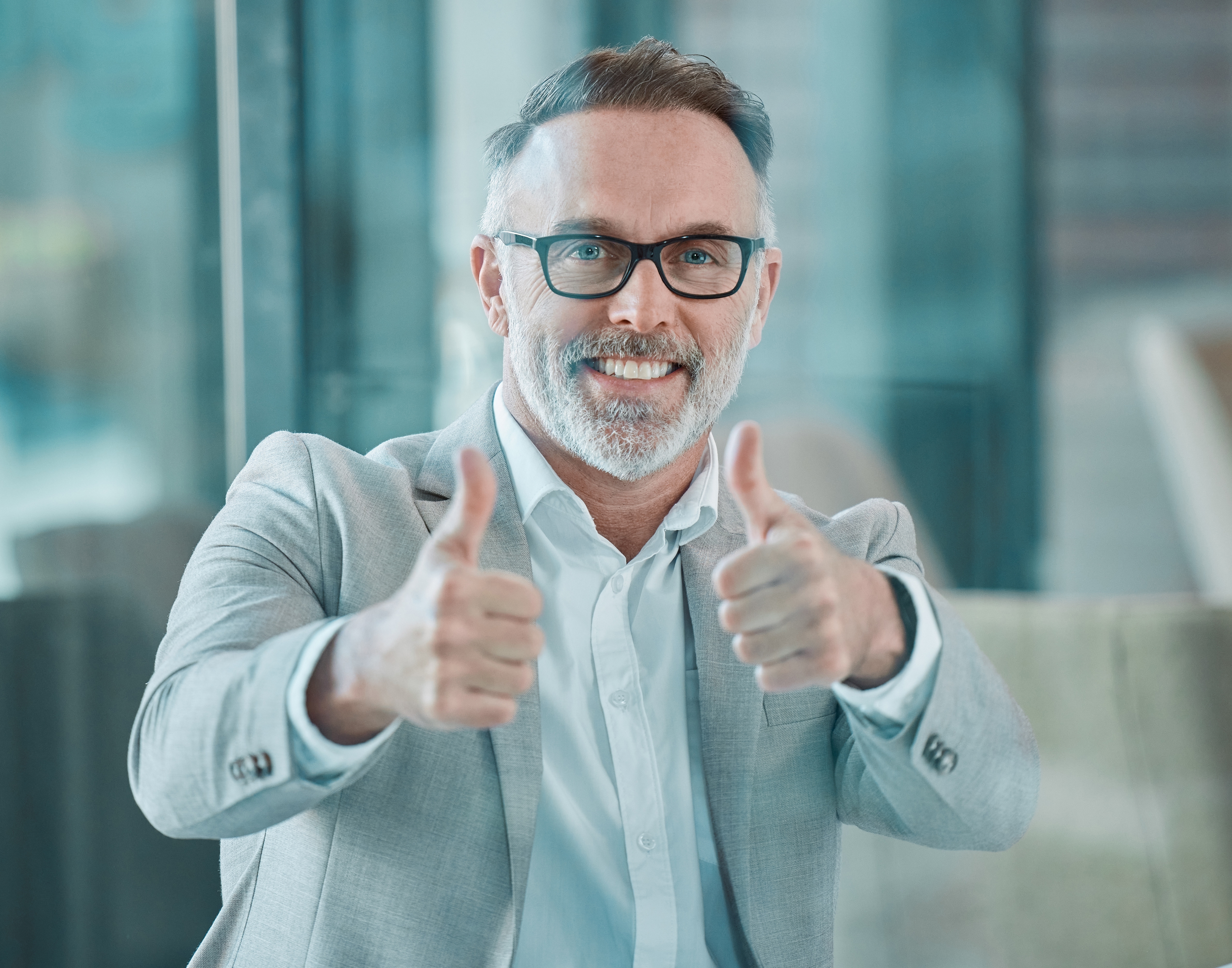 Medelålders man med glasögon och grå kavaj sitter inomhus, ler och visar tummen upp med båda händerna.