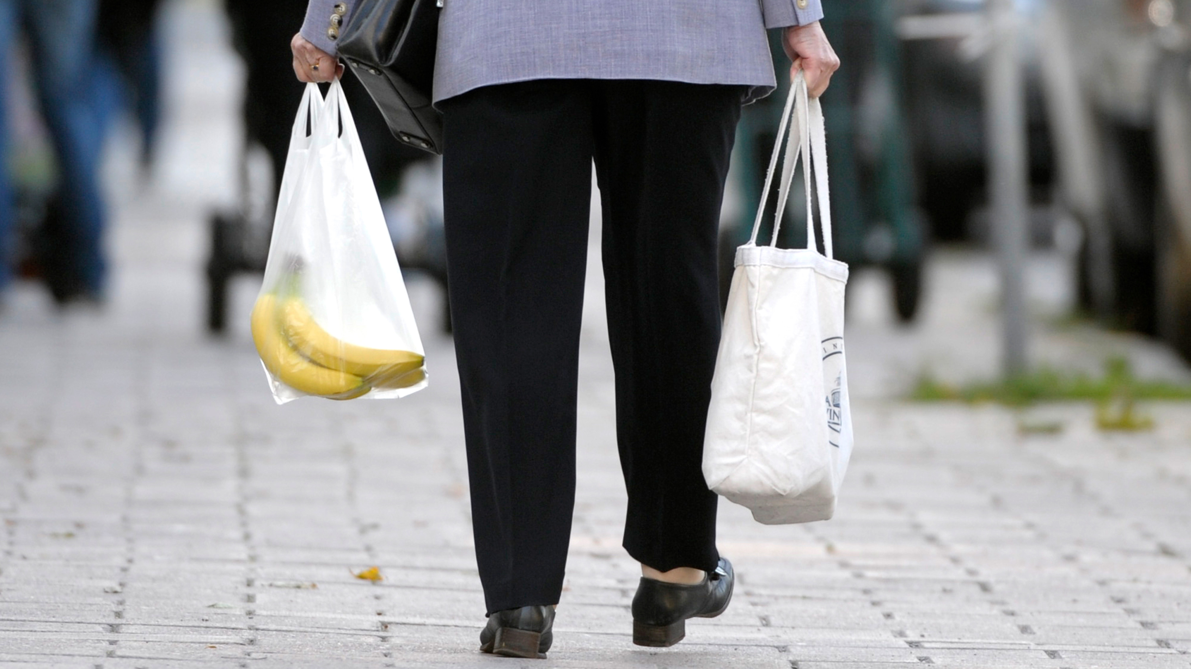 En person går på en trottoar och håller en plastpåse med bananer i ena handen och en återanvändbar vit kasse i den andra.