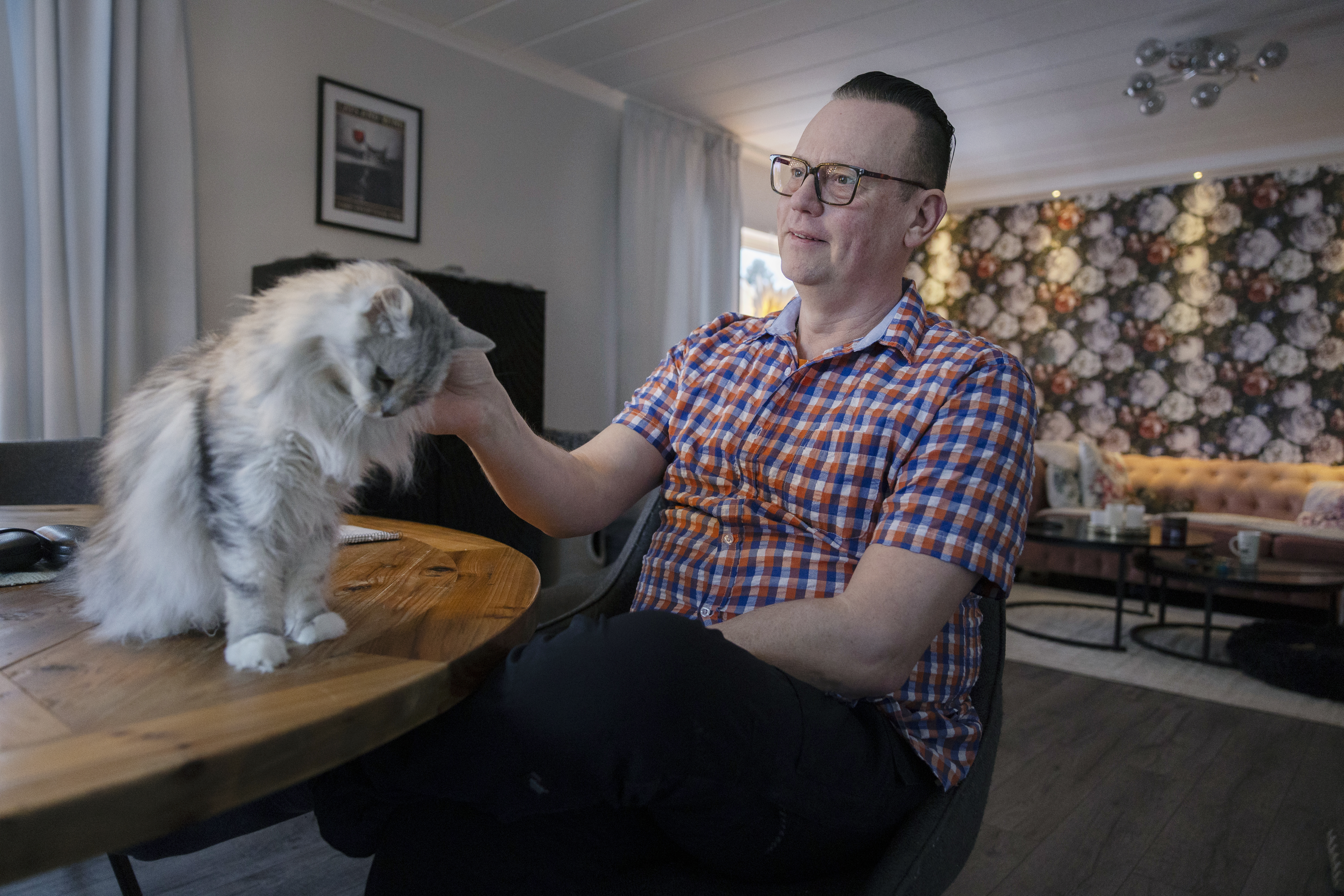 En person med glasögon och rutig skjorta sitter vid ett bord, klappar en långhårig katt i ett vardagsrum med blommiga tapeter och en soffa.