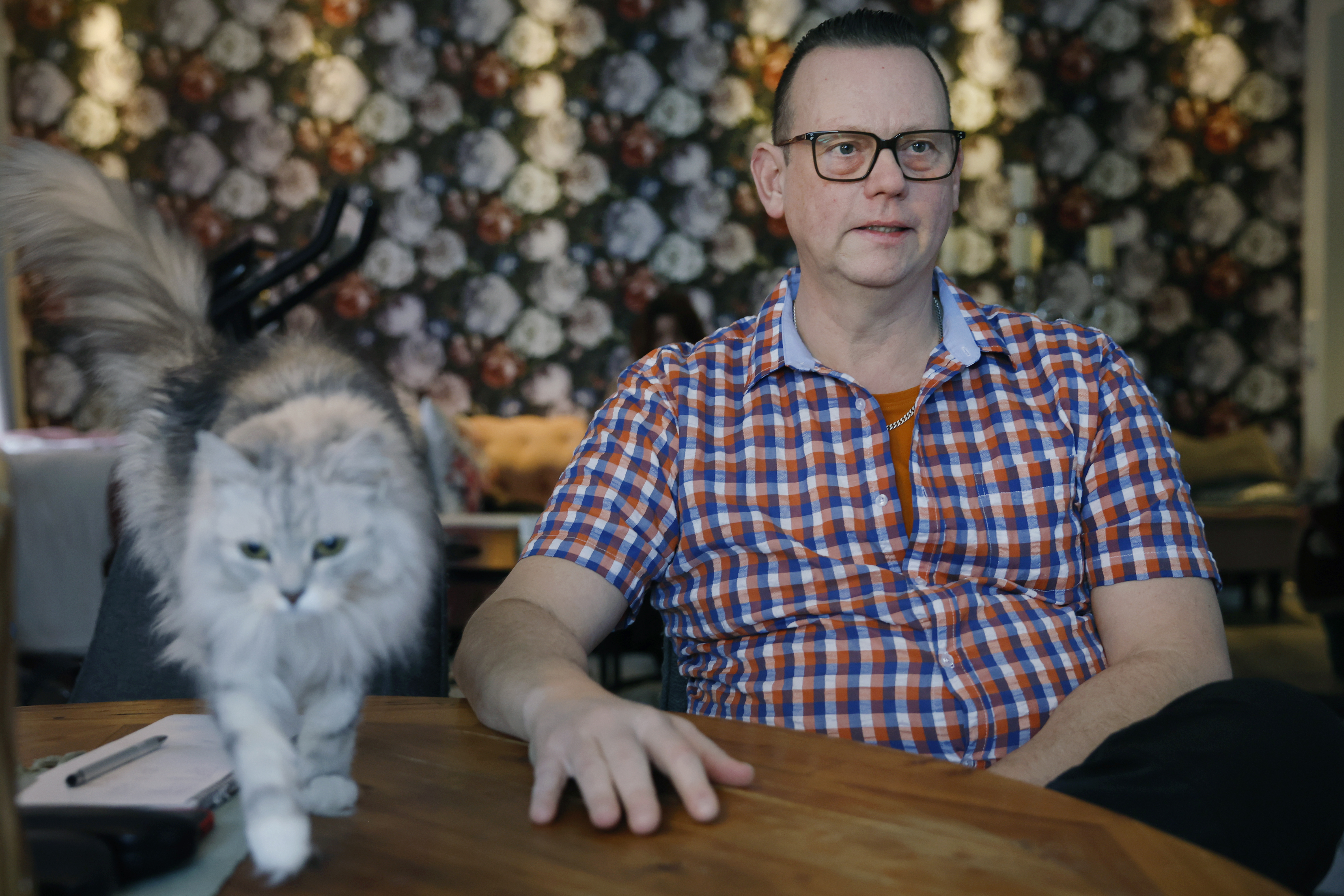 En man med glasögon och rutig skjorta sitter vid ett bord. En katt går bredvid. Bakgrunden har blommigt tapetmönster.