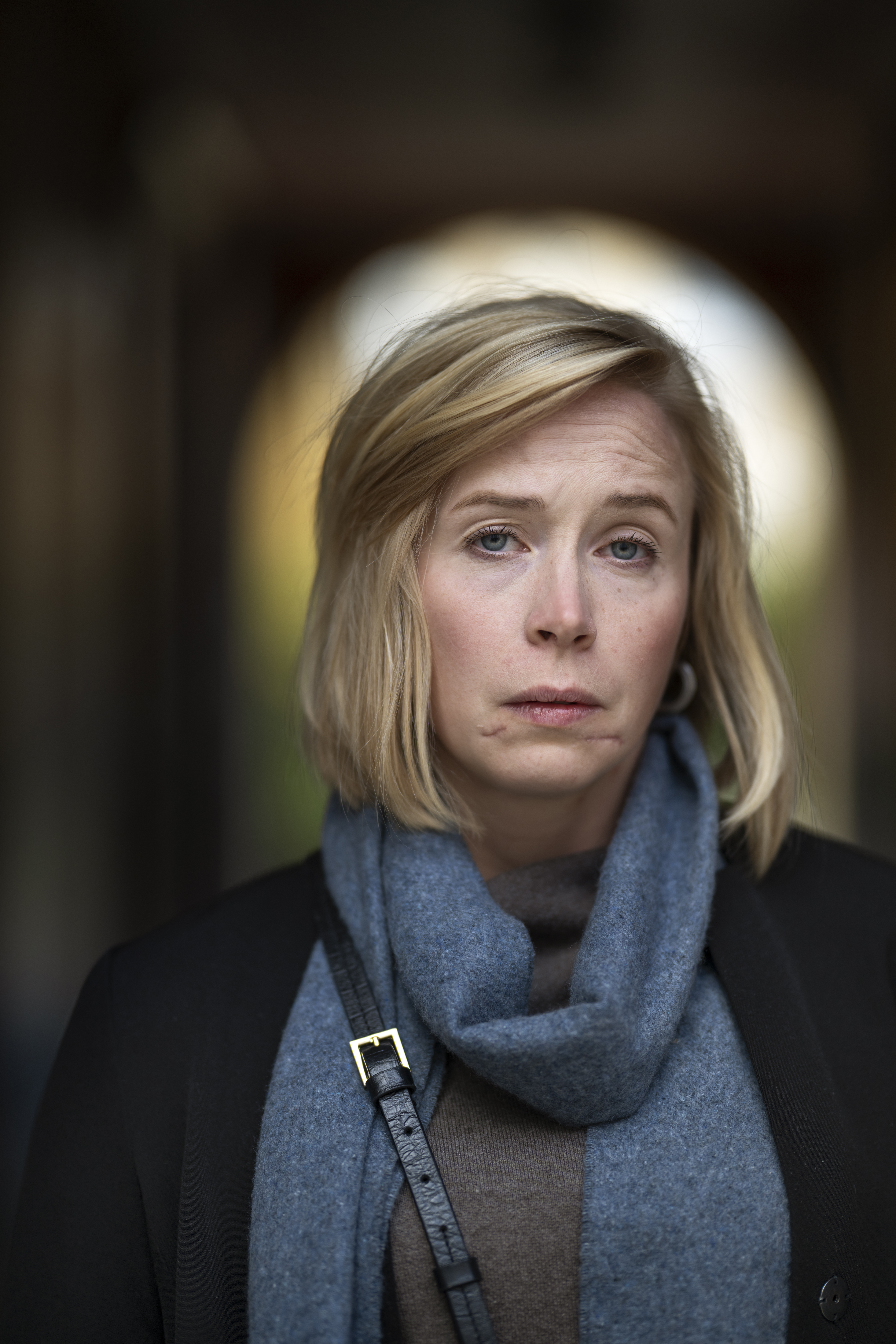 En kvinna med blont hår har en blå halsduk och mörk jacka, och tittar rakt in i kameran med neutral min.