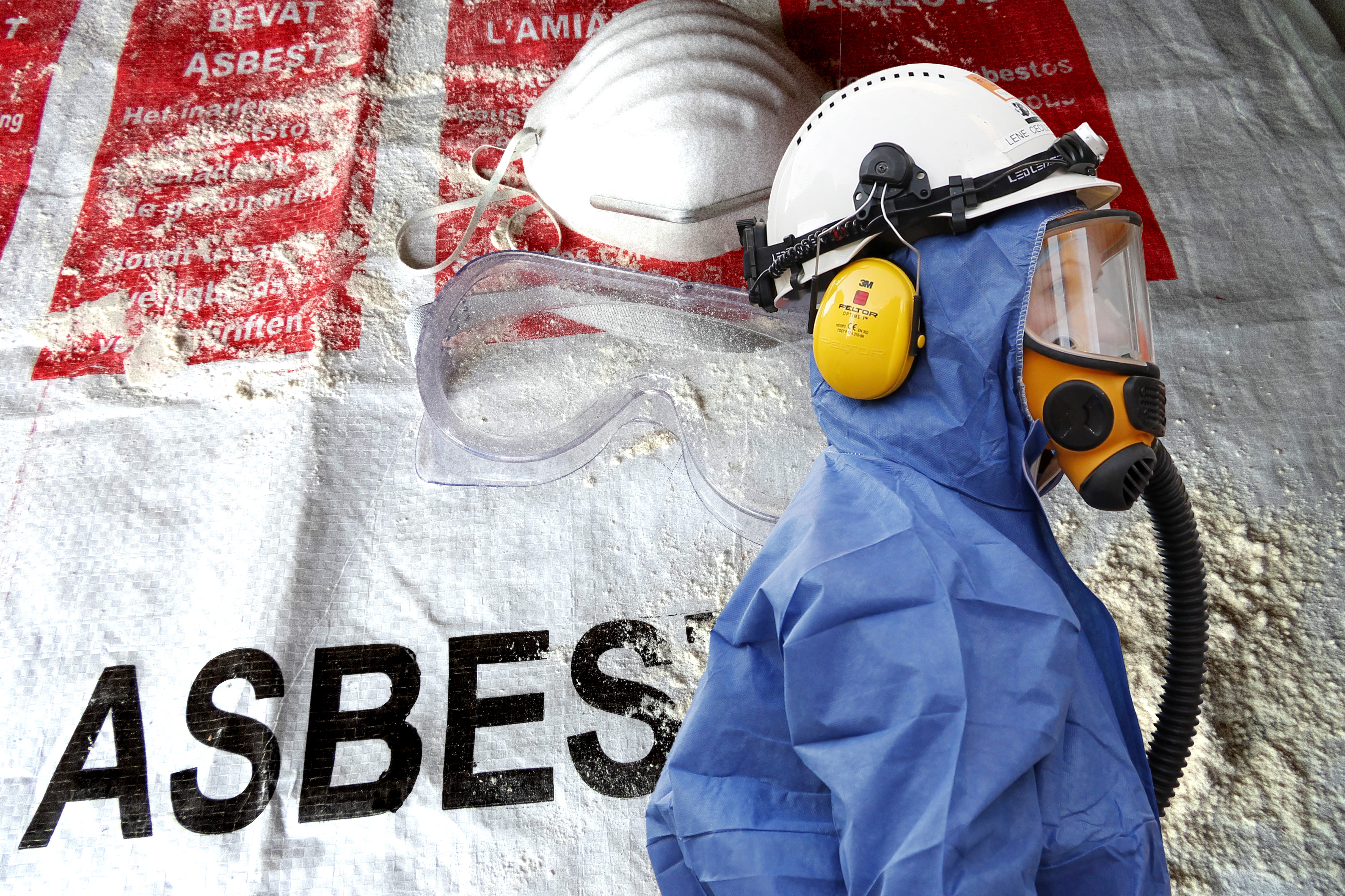 Skyddsutrustning bestående av mask, skyddsglasögon och hörselskydd på en blå dräkt, placerad på ett väska märkt "ASBESTOS".