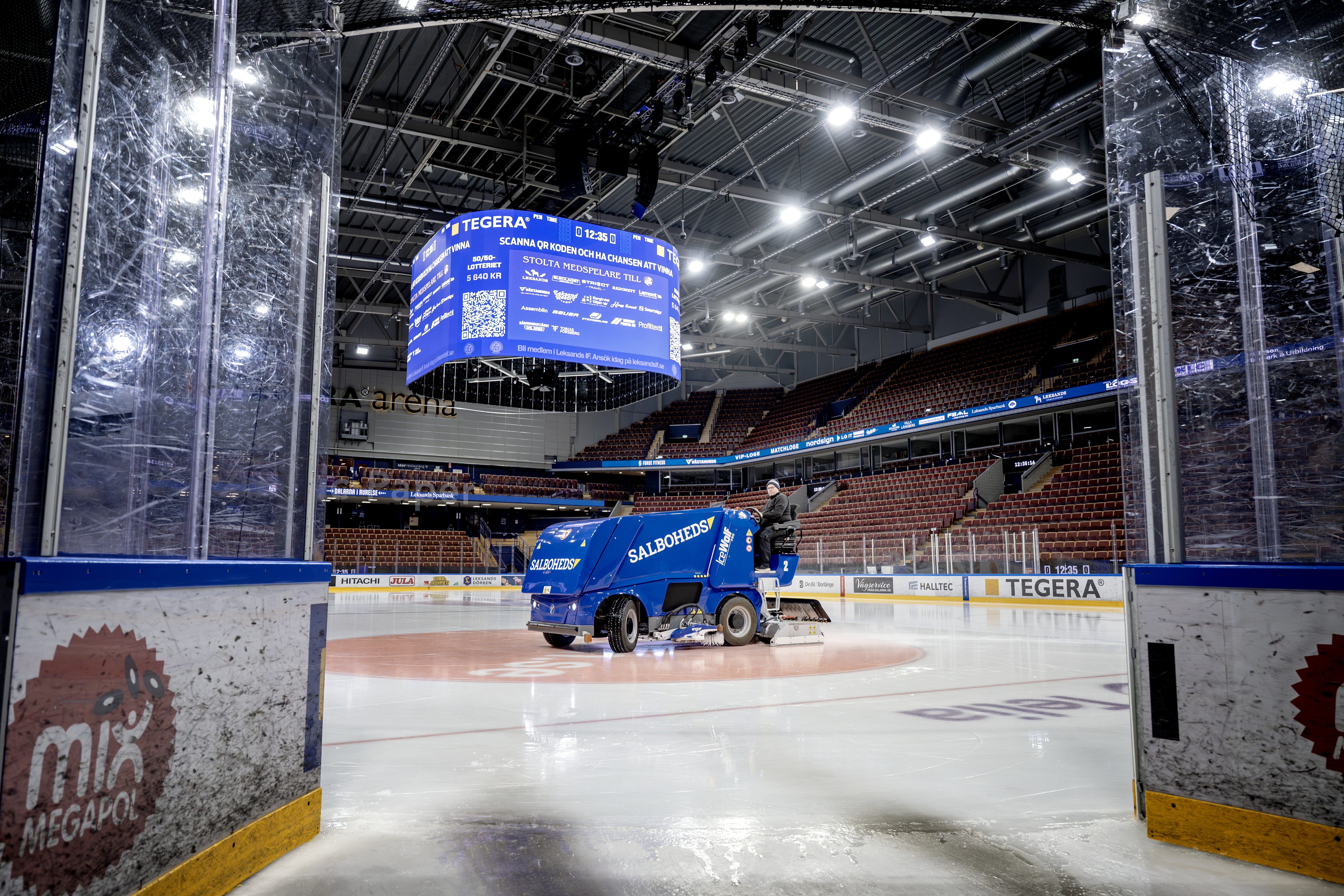 En ismaskin ute på isen i en tom ishall. Läktare och en digital resultattavla syns i bakgrunden.