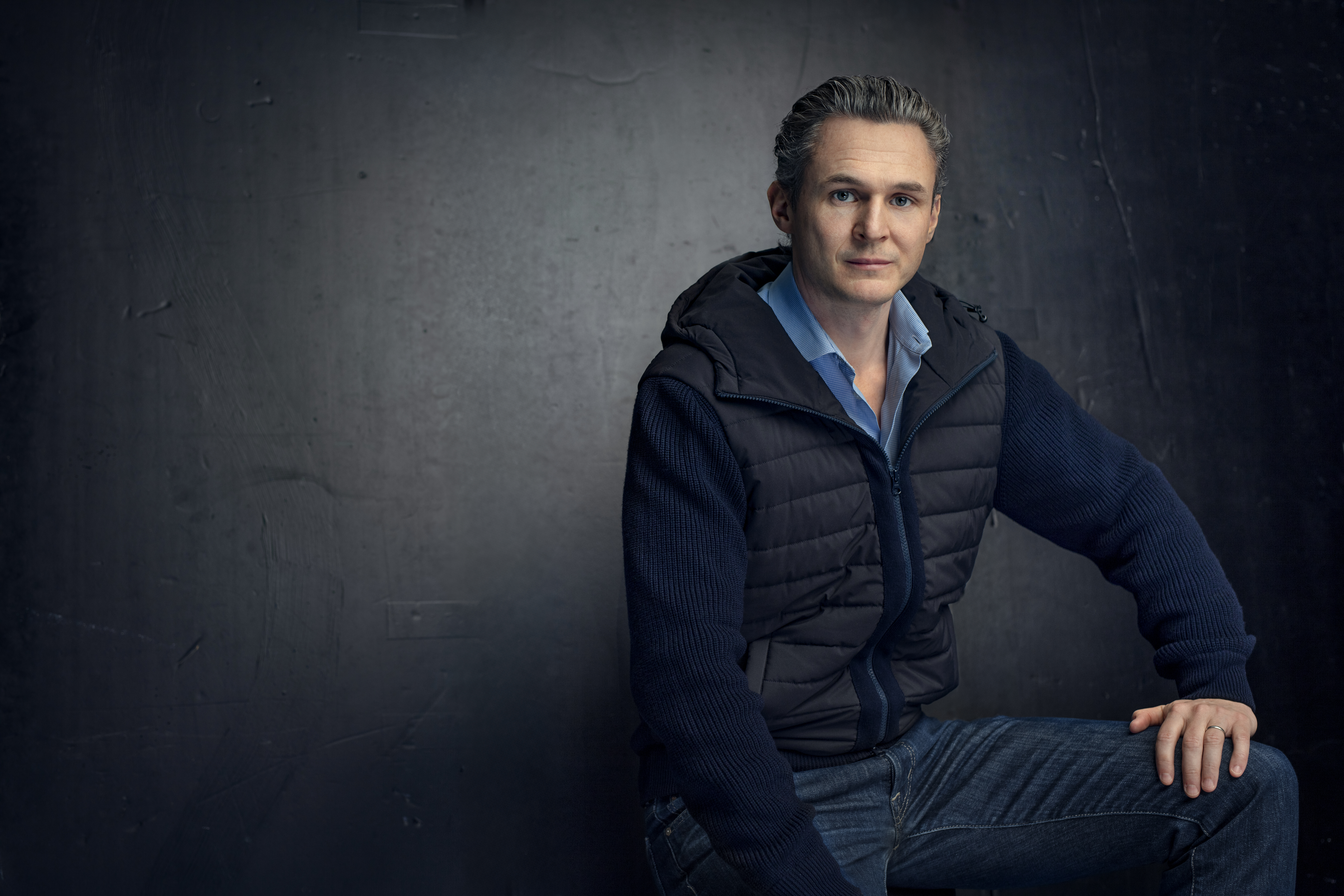 En man med kort grått hår sitter på en pall mot en mörk bakgrund, klädd i blå skjorta, mörk tröja och jeans.