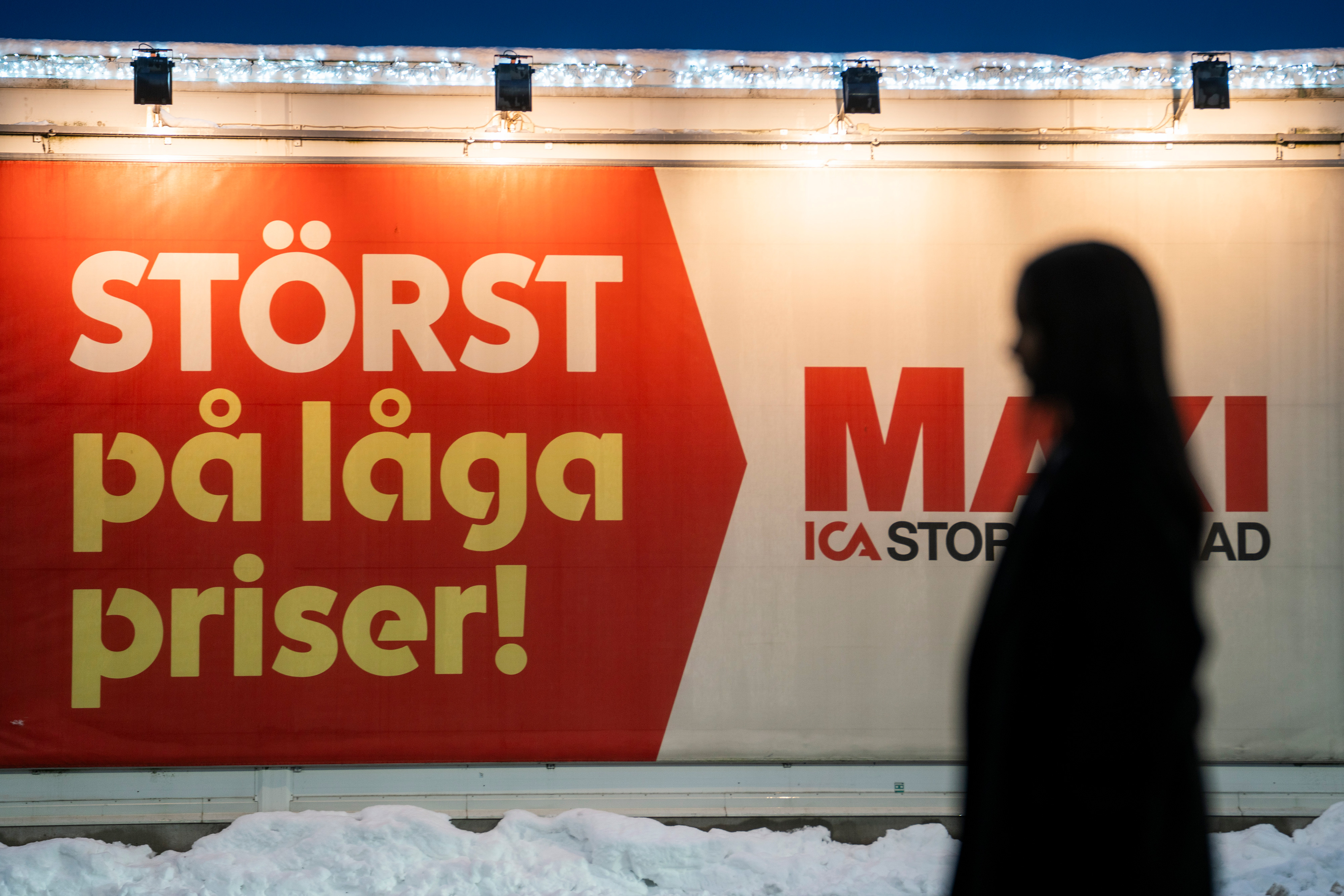 En person i silhuett går förbi en skylt på svenska som annonserar låga priser vid Maxi ICA, med snö på marken.