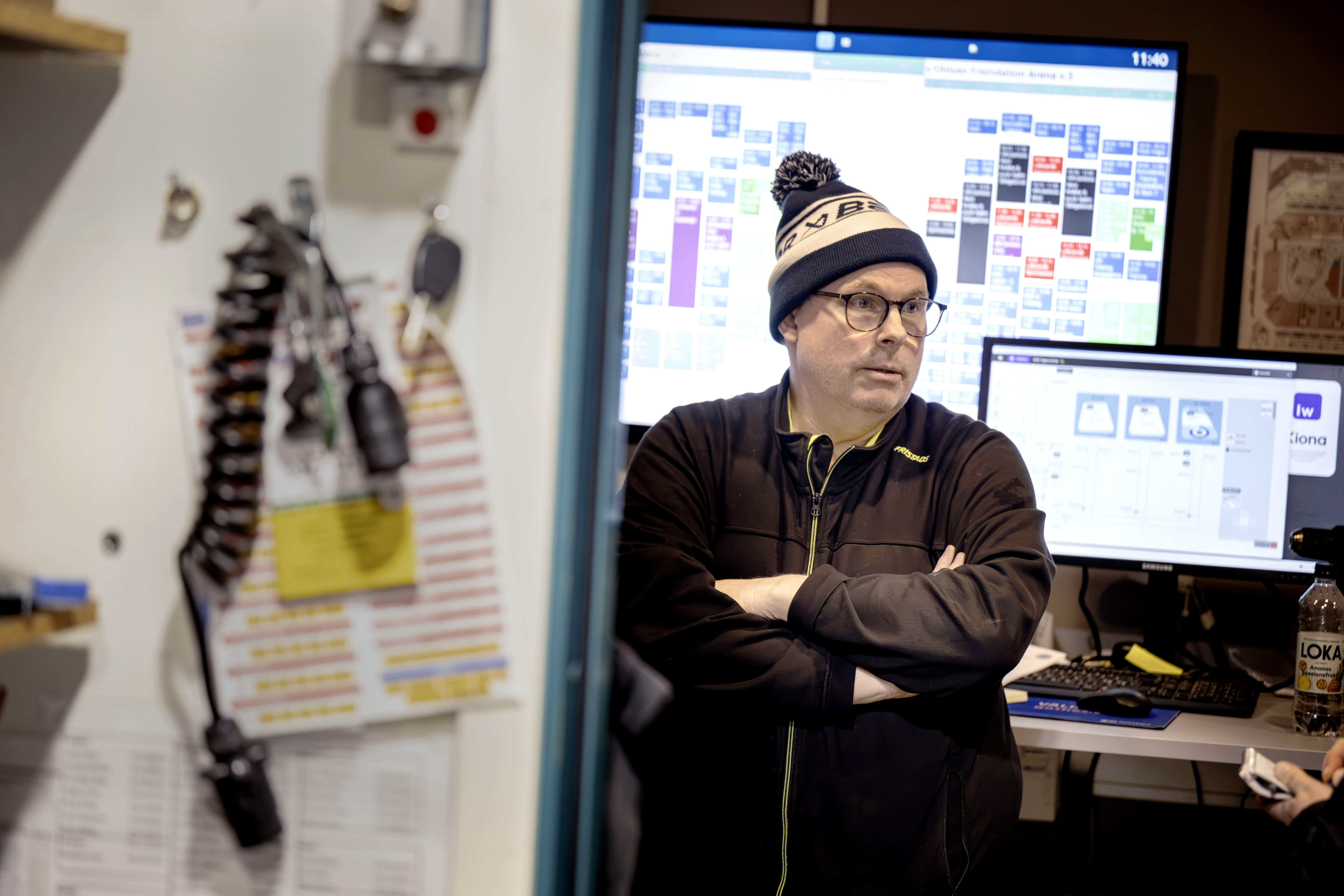 Stefan Bäcker med glasögon, mössa och jacka står med armarna i kors i öppningen till et rum där datorskärmar och utrustning syns.