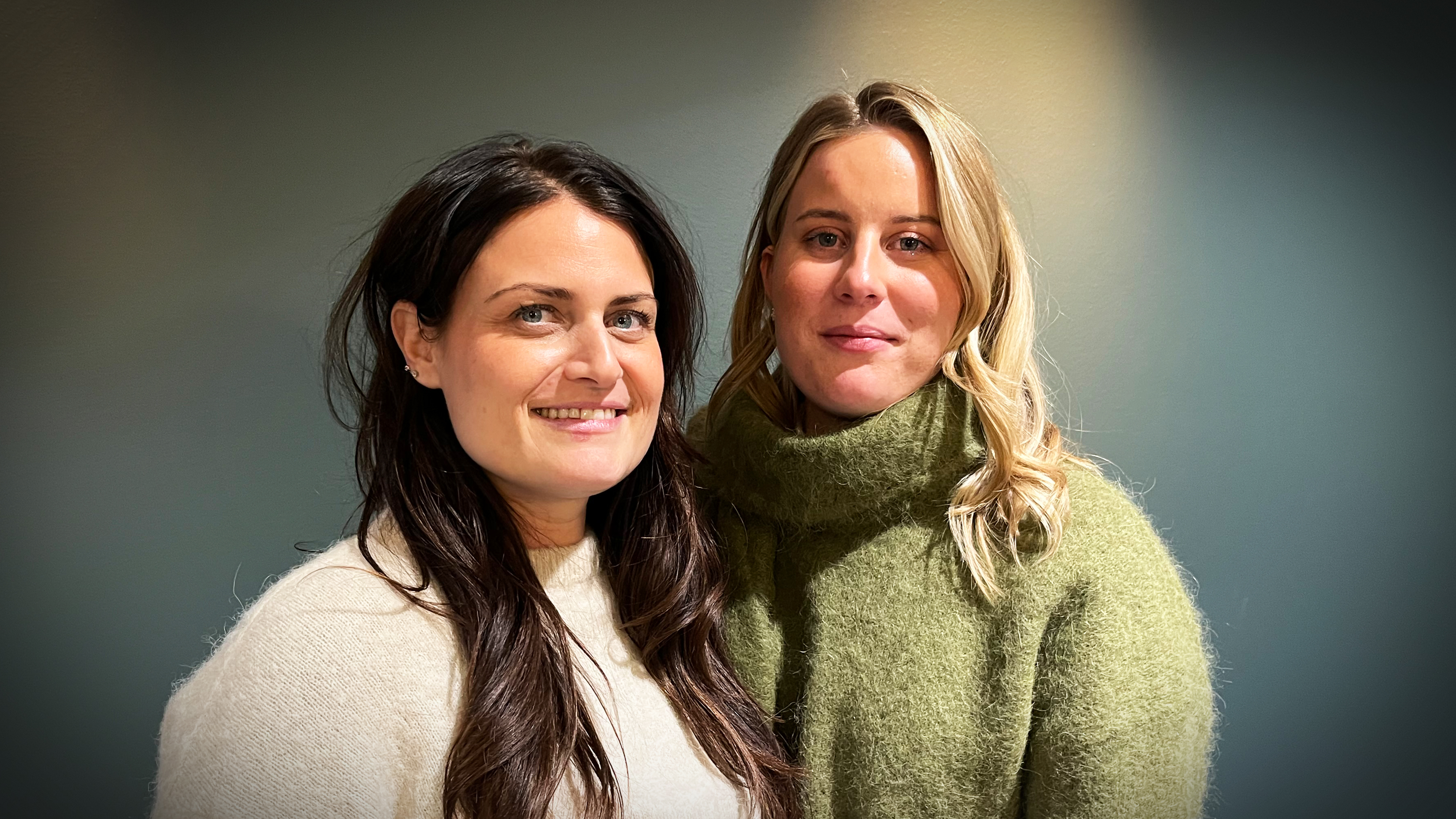 Två kvinnor med långt hår står bredvid varandra framför en blågrå vägg, båda har tröjor och tittar mot kameran.