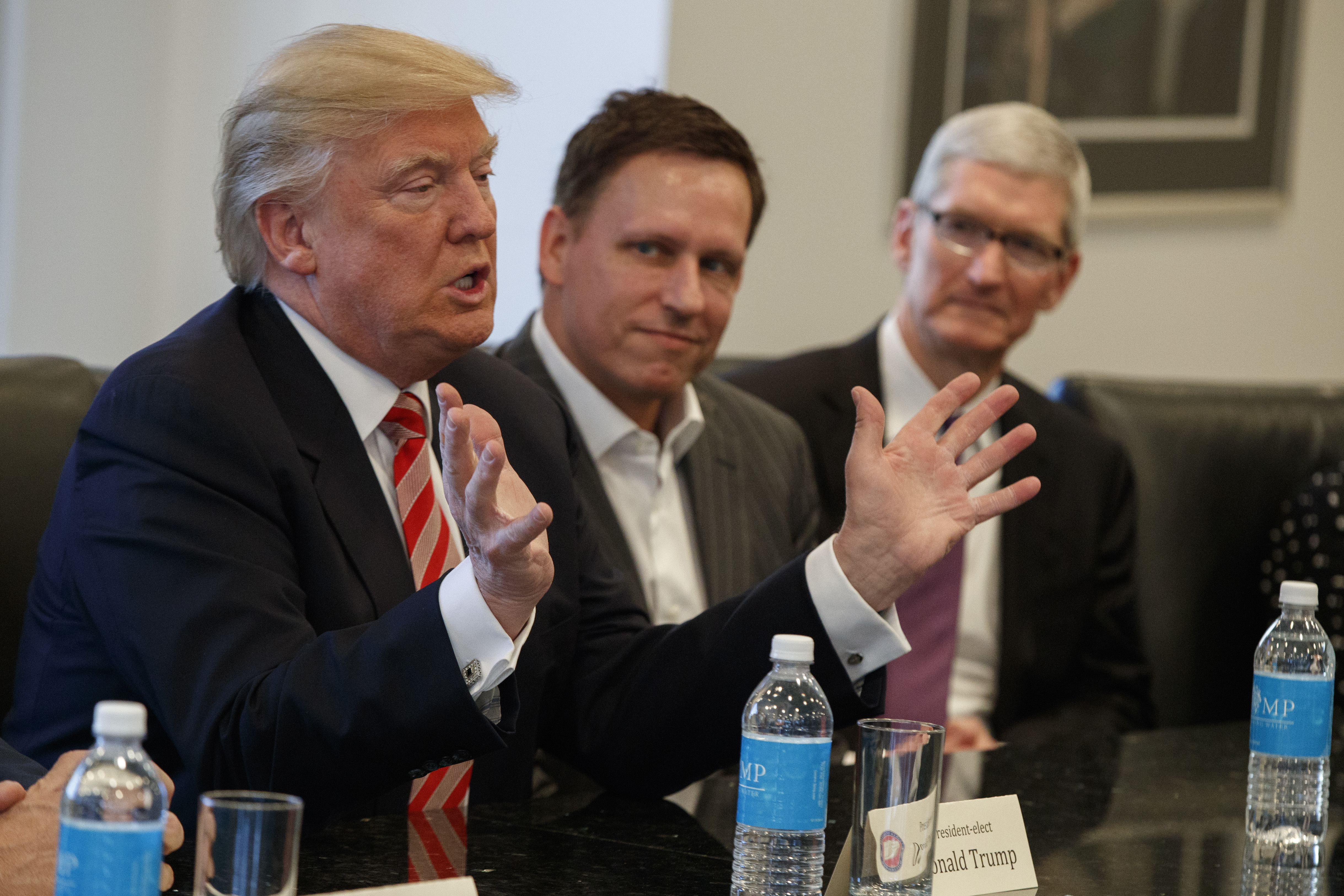 Donald Trump talar vid ett konferensbord med flera män, däribland Peter Thiel och Tim Cook. Flaskor och namnskyltar finns på bordet.